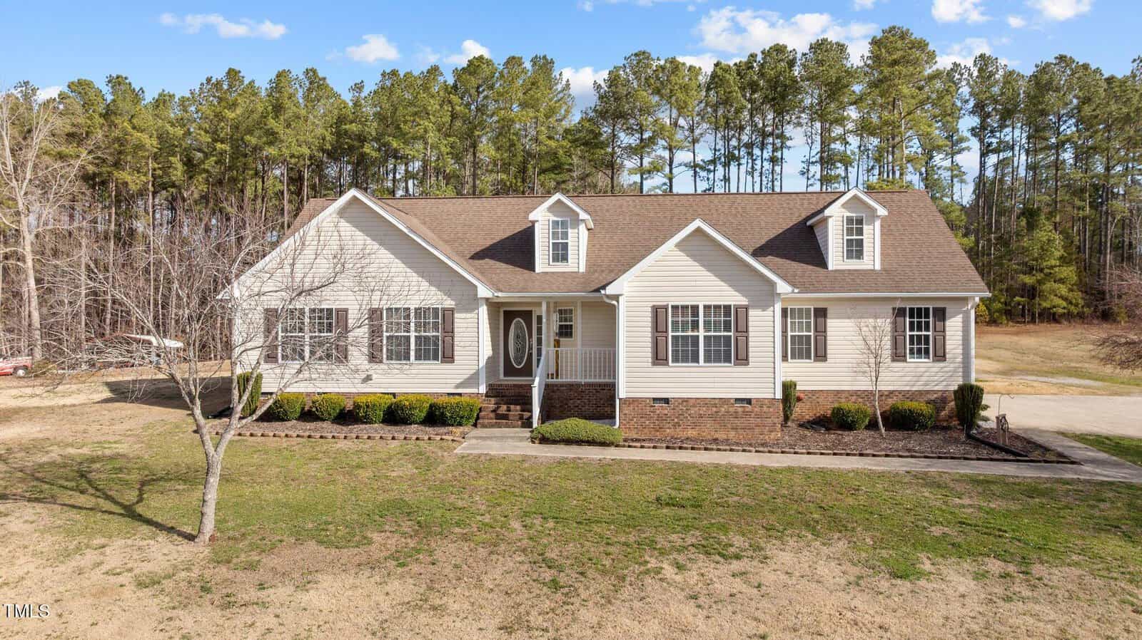 A single-story house at 1031 Franklin Lane is pictured, showcasing beige siding and a brown shingled roof. It features white trim, a central front door with sidelights, and small shrubs along the front. The home in Henderson is set against tall pine trees and a blue sky.