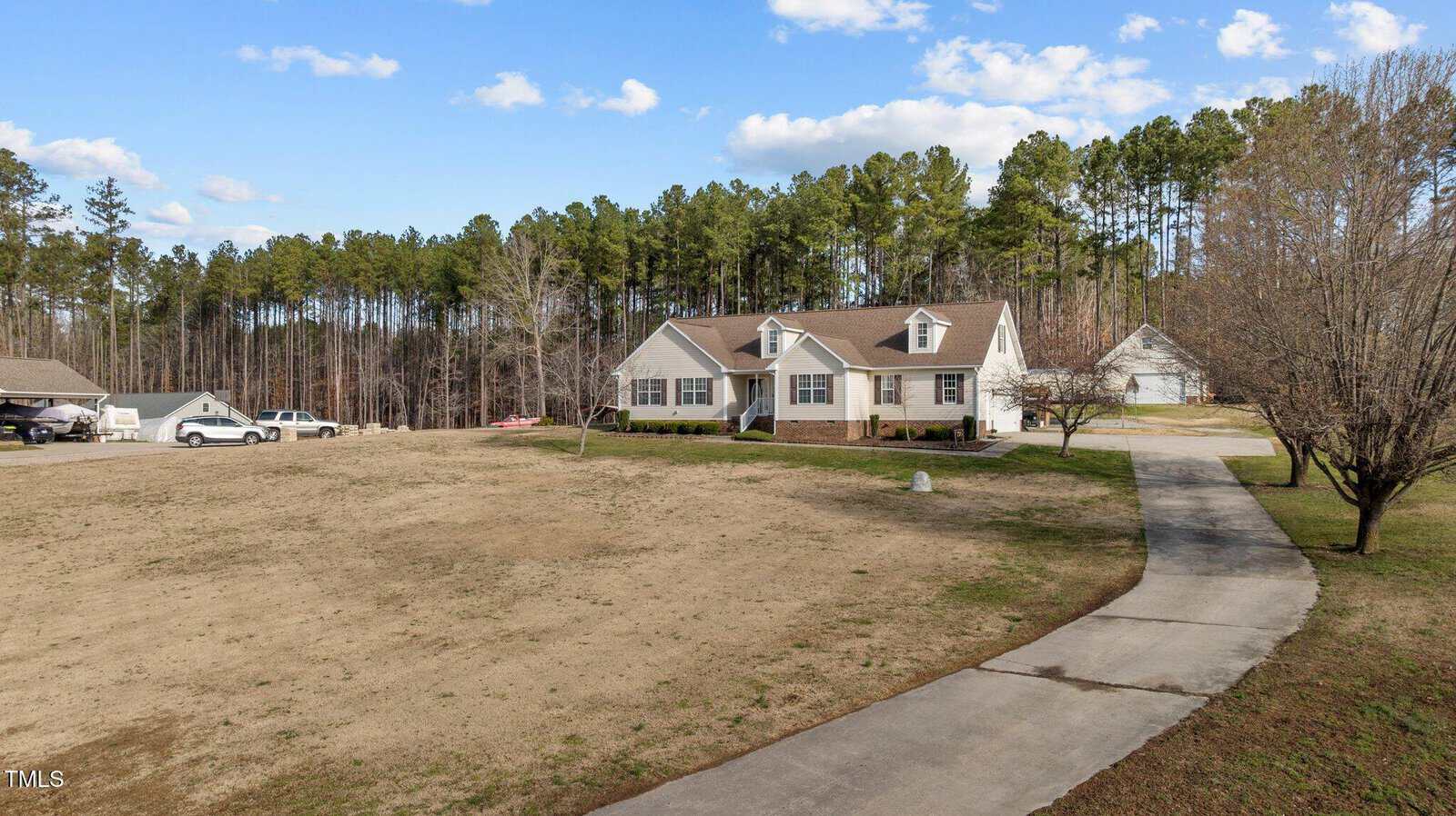 A house with a beige exterior sits on a large plot of land, featuring a long driveway leading to the property. The surrounding area is grassy with a few trees and a wooded area in the background. Several vehicles are parked near the house.