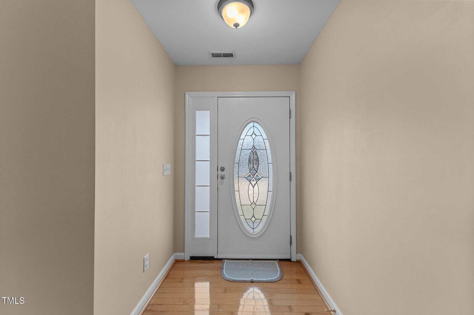 A hallway with light beige walls and wooden flooring leads to a white front door with an oval, decorative glass panel. A ceiling light fixture illuminates the space, and a small rectangular rug lies in front of the door.