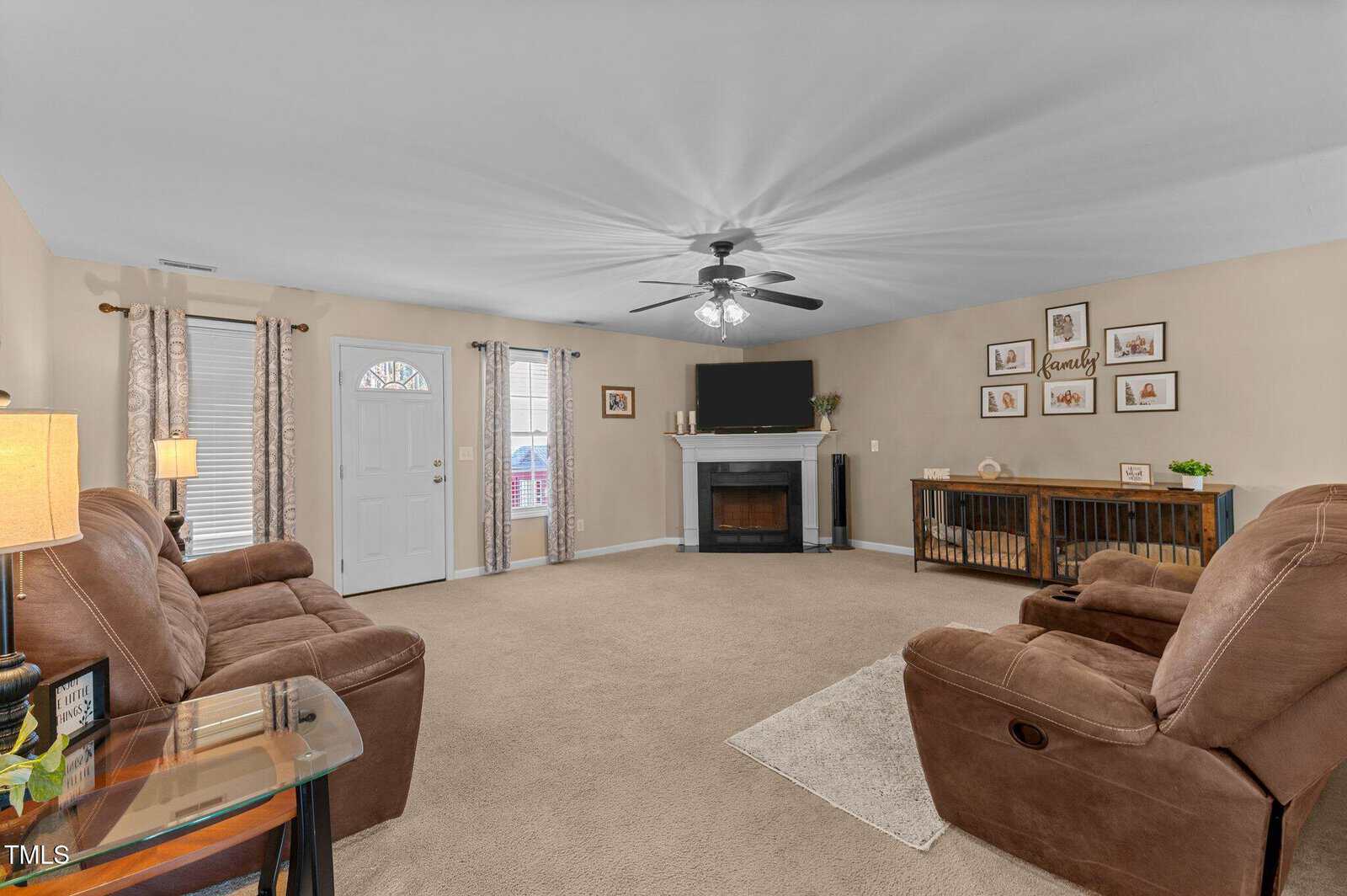 Spacious living room with beige carpet and light brown walls. Features brown reclining sofas facing a fireplace with a TV above it. A ceiling fan hangs above, and three windows and a door provide natural light. Decorative items and wall art adorn the space.