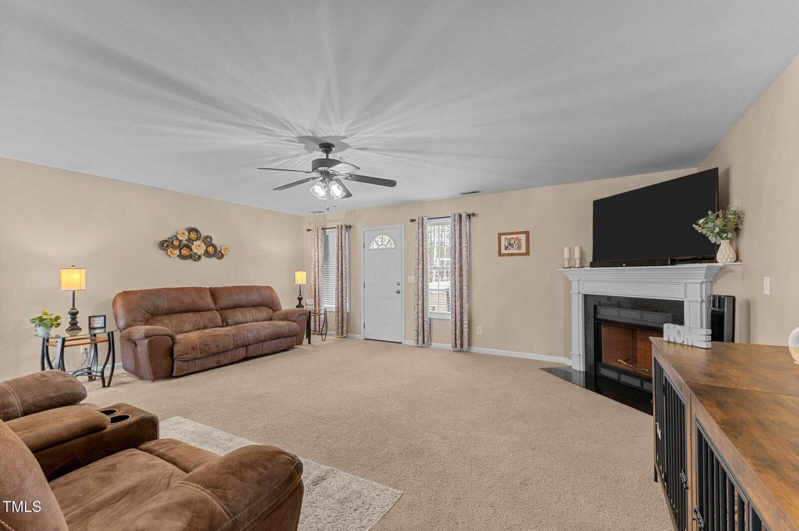 A spacious living room with beige walls and carpet features a brown reclining sofa and chair. A fireplace with a TV above it is on the right. There's a ceiling fan with lights, two small windows, and a white front door in the background.