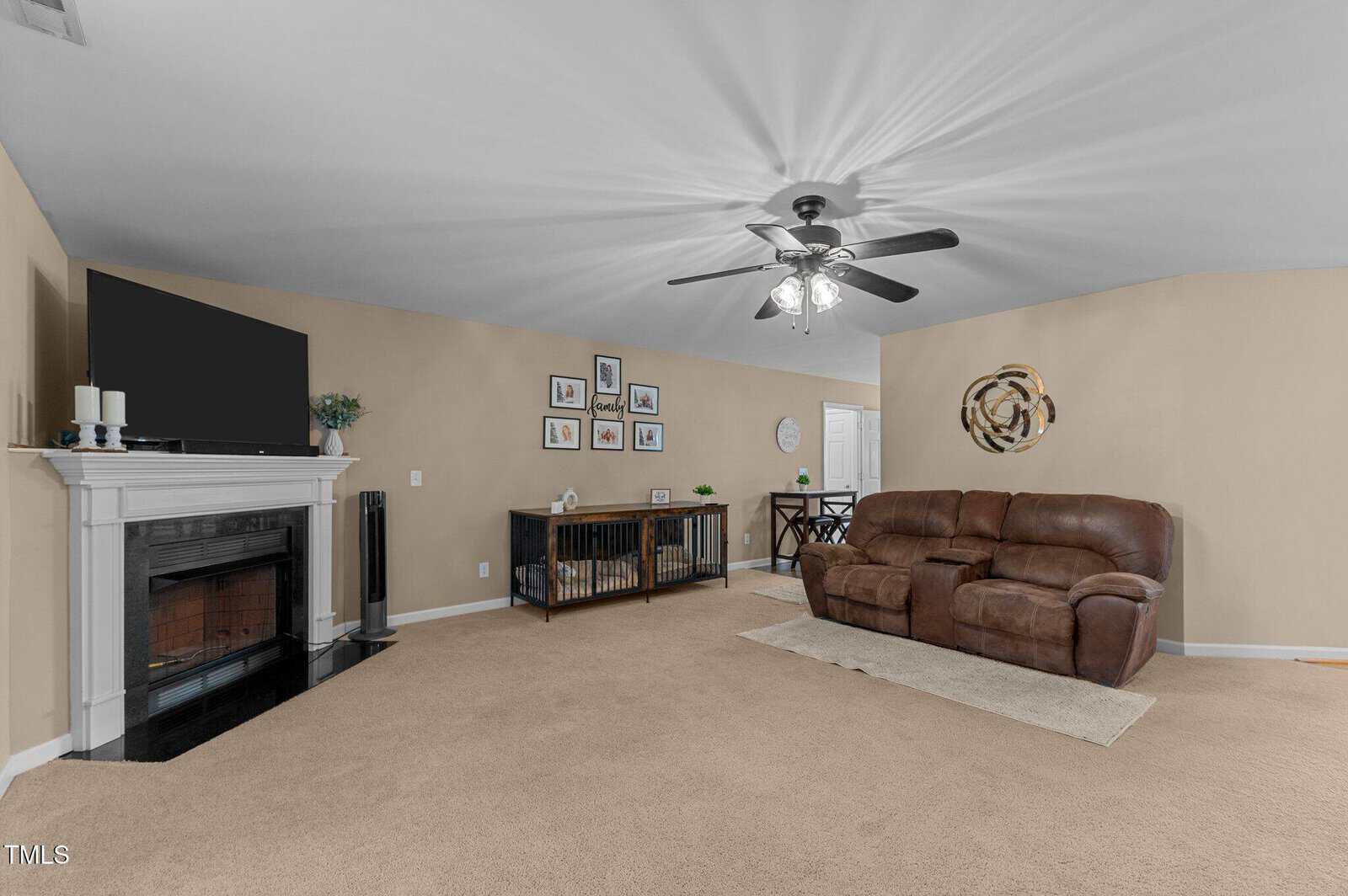 A living room with beige walls and carpet features a dark wood fireplace with a TV on top. There is a brown leather sofa, a sideboard with picture frames above it, and a ceiling fan with lights. A small dining area is visible in the background.