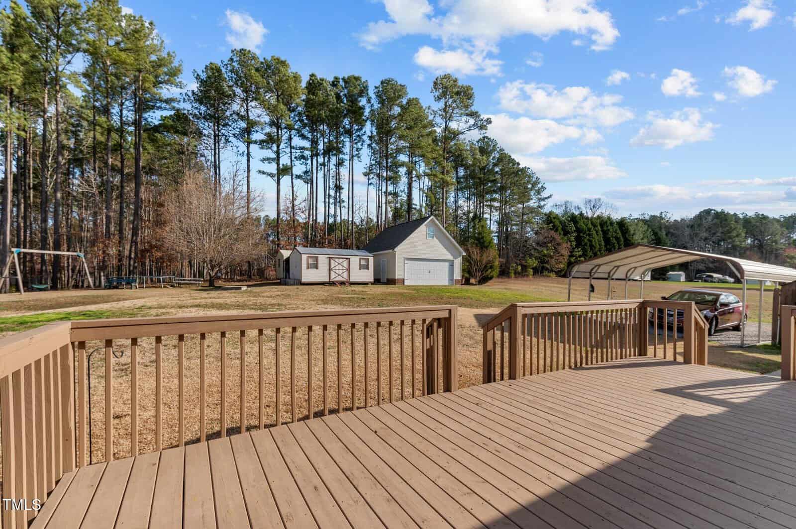 A wooden deck overlooks a spacious backyard with a shed and a garage. Tall trees line the perimeter. A swing set is on the left, and a carport shelters a red vehicle on the right. The sky is clear with scattered clouds.