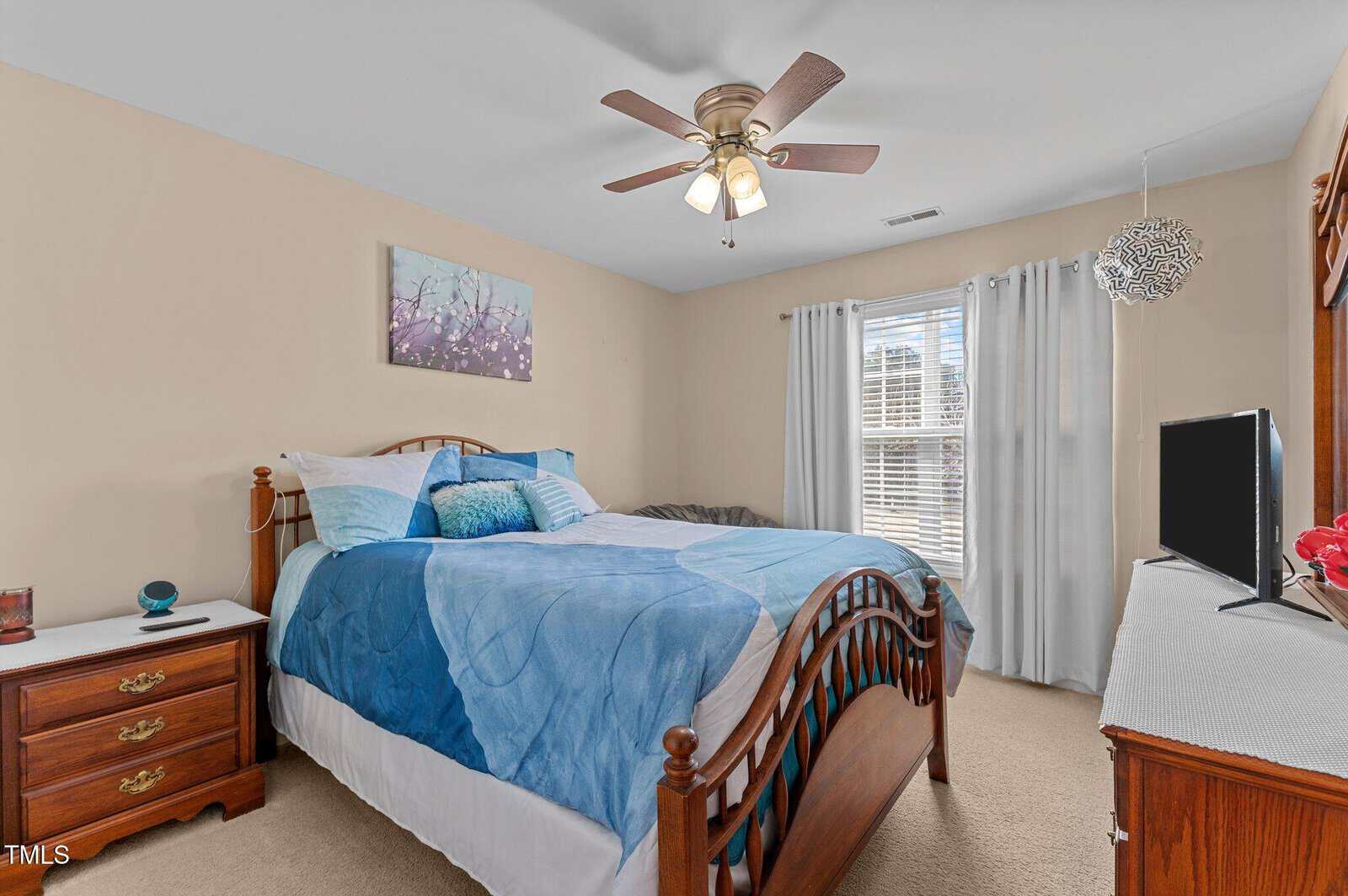 A bedroom with a wooden bed frame and blue bedding. There's a wooden nightstand with a lamp on the left and a dresser with a TV on the right. A ceiling fan is above, and a window with white curtains lets in natural light. A painting hangs on the wall.