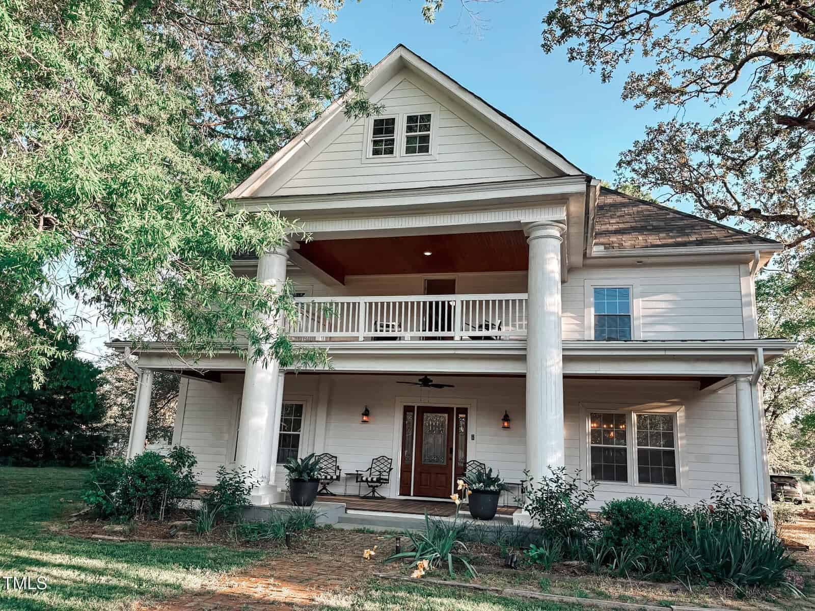 A two-story white house with large columns, a covered front porch, an upper balcony, and a dark wooden front door. The yard has green plants and trees, and there is a ceiling fan on the porch.