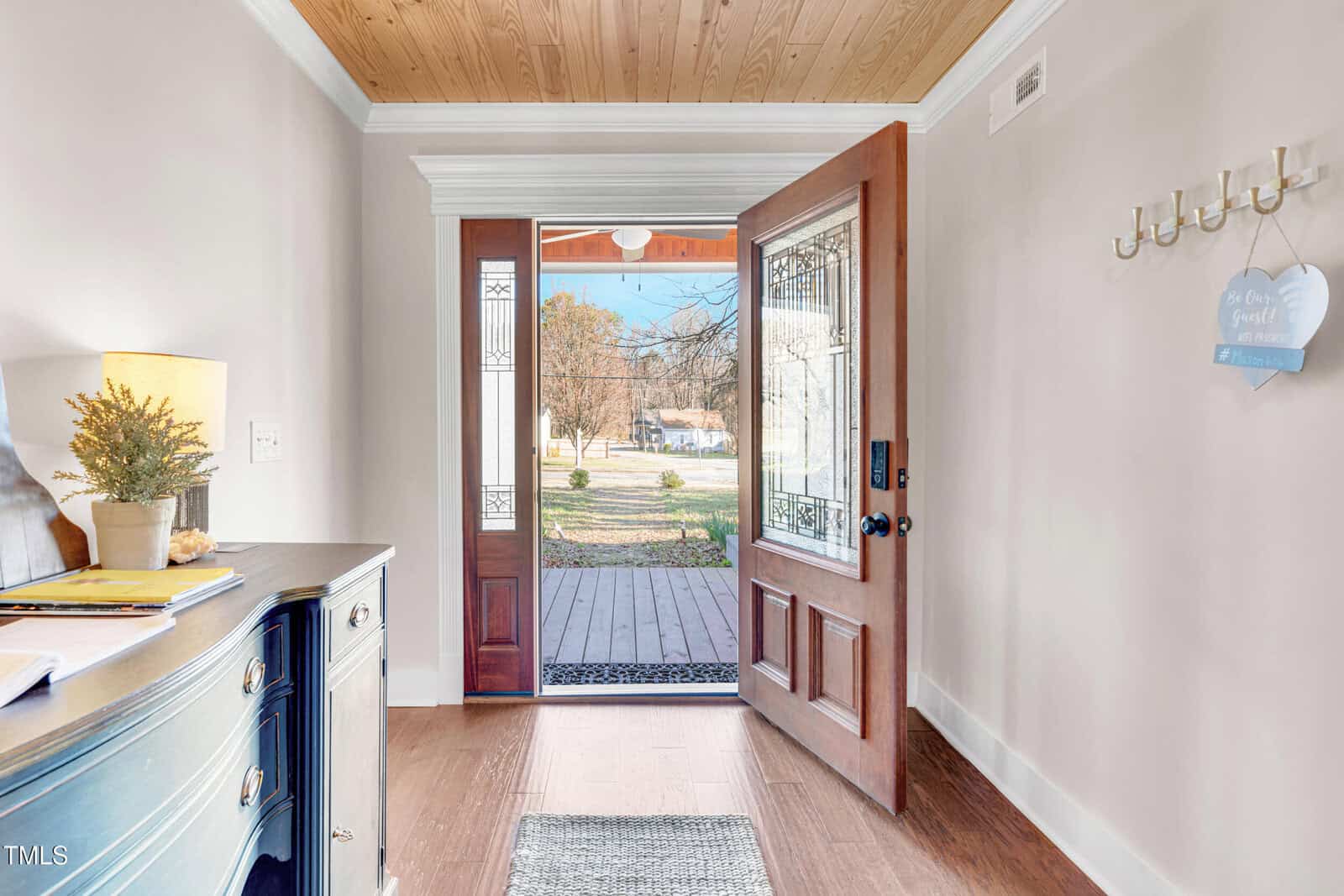 A front entryway with a wooden door open to a view of the porch and yard outside. The area includes a dark wood cabinet, a small potted plant, and wall hooks with a sign. Sunlight brightens the space.
