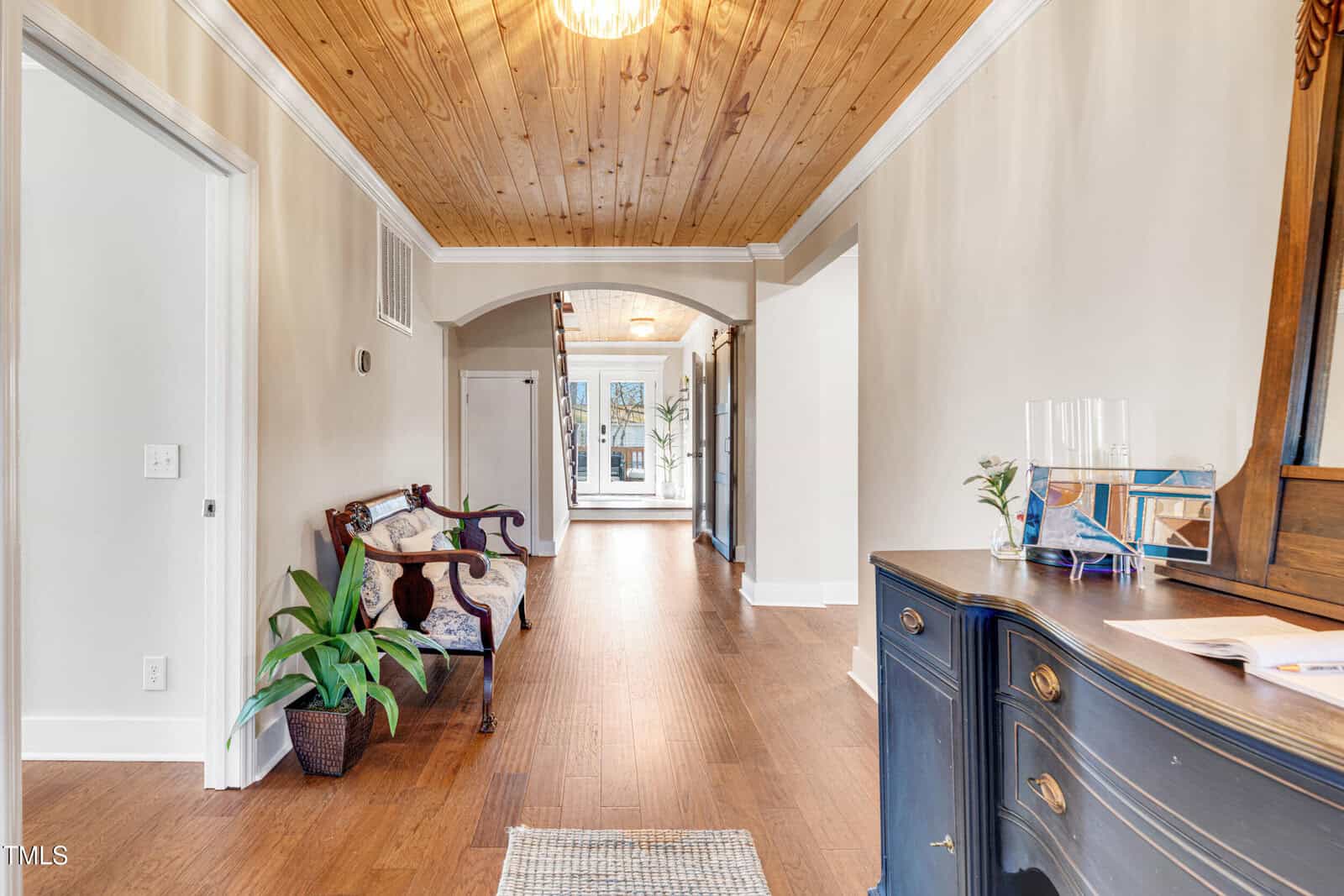 A hallway with wood flooring and a wood-paneled ceiling, featuring a dark wood bench with cushions, potted plants, a blue dresser with a mirror, and an arched entry leading to a room with glass doors.