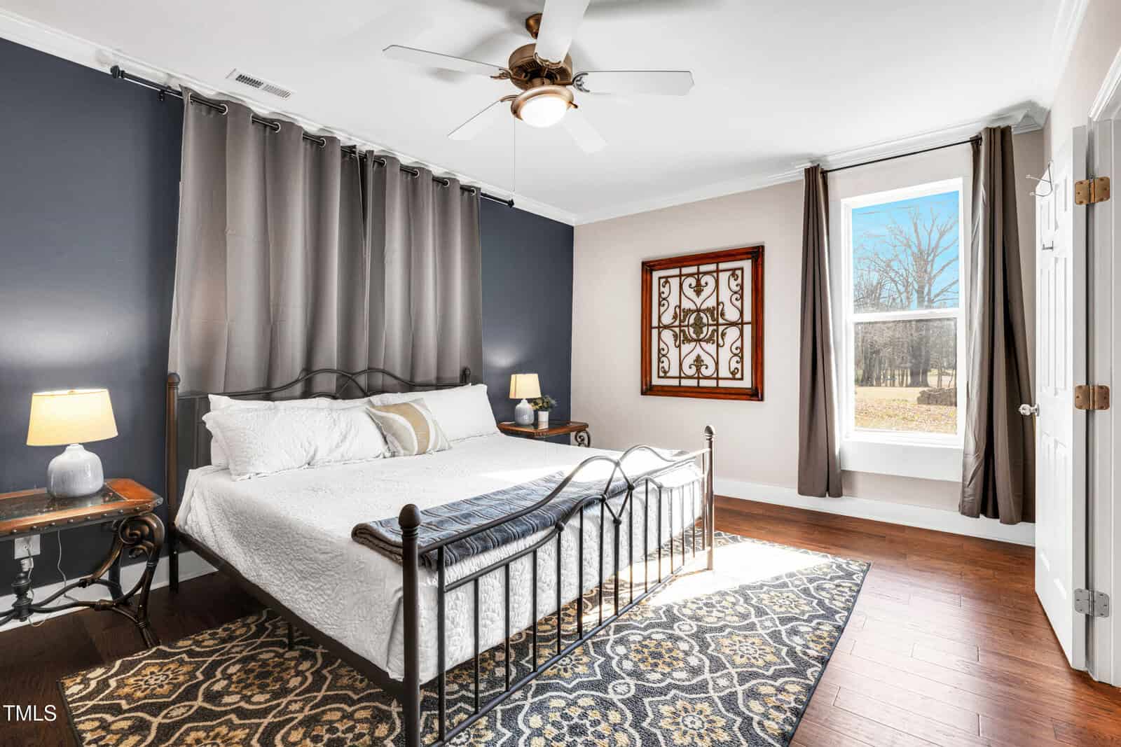 A bedroom with a metal bed frame, white bedding, two bedside tables with lamps, a ceiling fan, patterned rug, dark blue accent wall with curtains, framed art, and a large window showing an outdoor view.