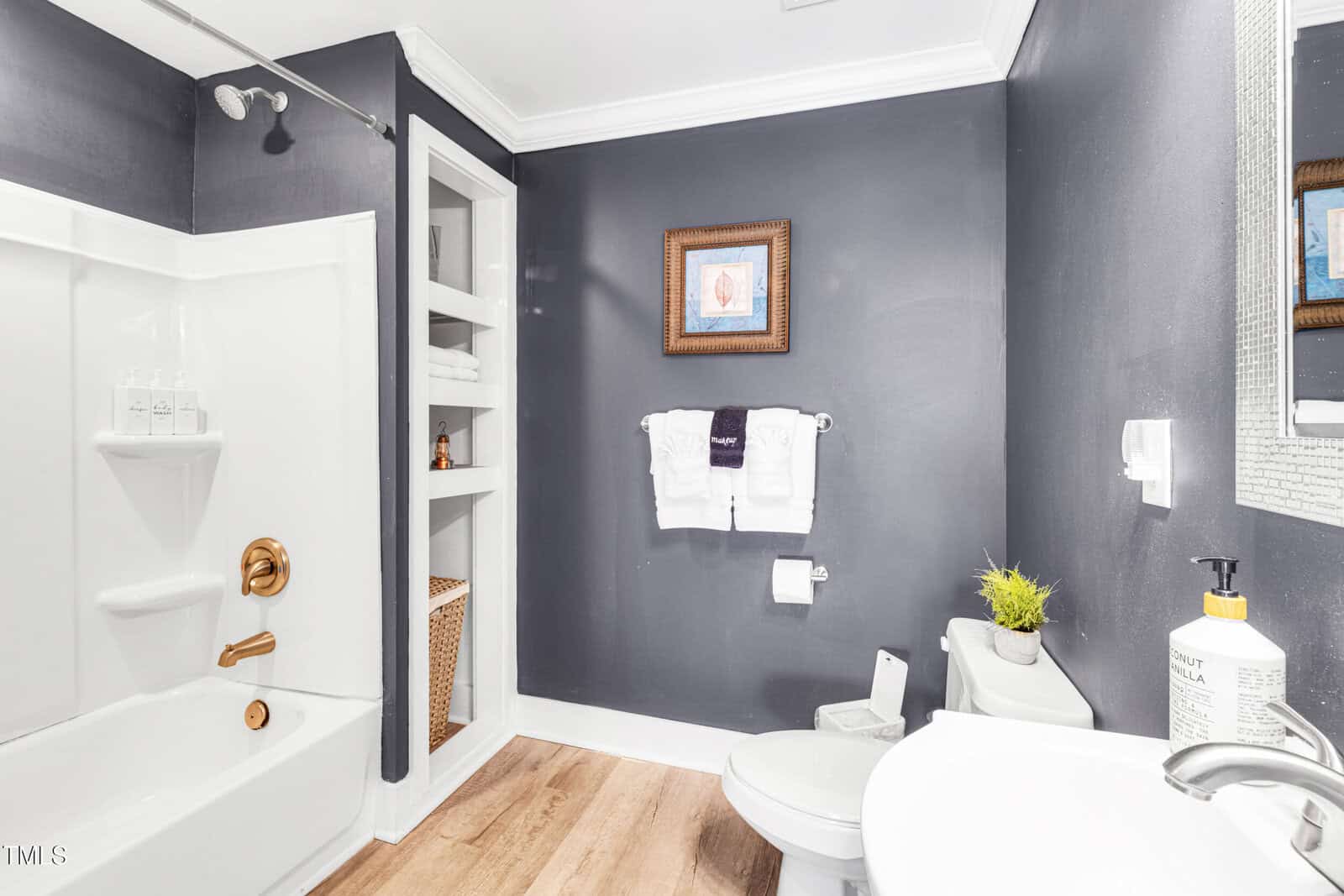 A bathroom with dark gray walls, a bathtub with gold fixtures, built-in shelves with towels and baskets, a toilet, a sink with soap and a plant, and a framed picture above the towel rack.