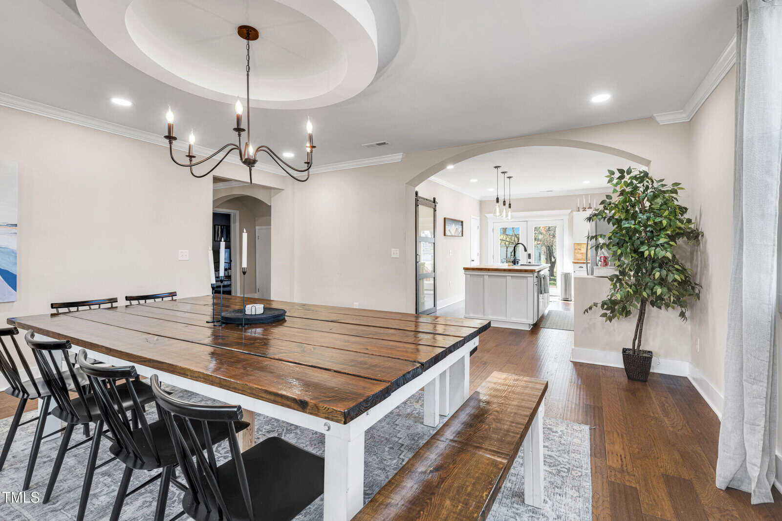Spacious dining room with a large wooden table, black chairs, and bench. Chandelier above the table, potted plant near the window, and arched entryway leading to a bright, modern kitchen. Hardwood floors throughout.