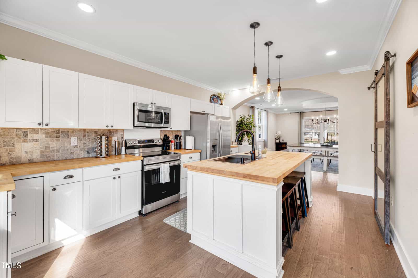 Modern kitchen with white cabinets, wood countertops, stainless steel appliances, center island with stools, pendant lights, and a backsplash with tan tile. Dining area with chandelier visible in the background.