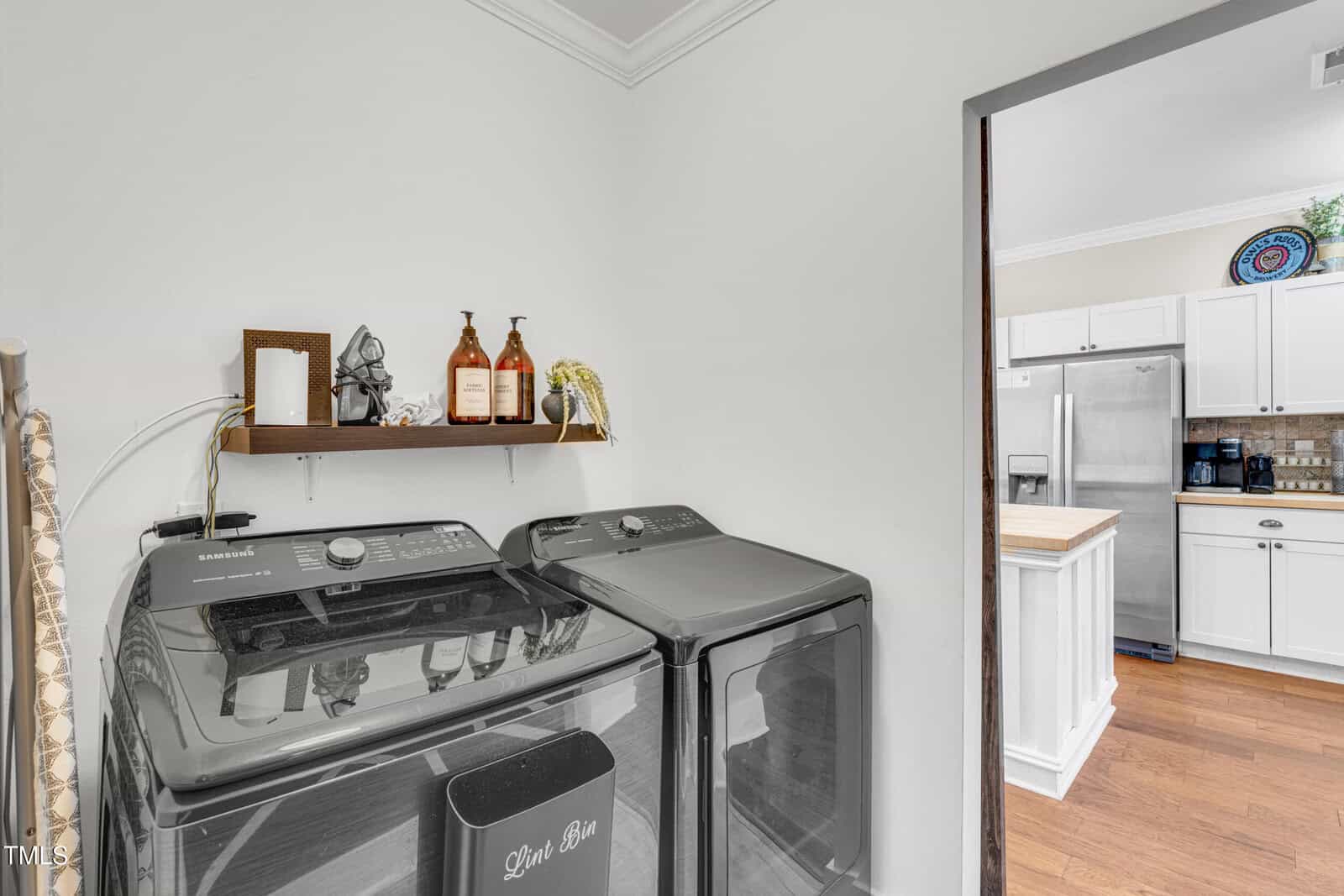 A laundry room with a black washer and dryer, a “Dark Bin” basket, a floating shelf holding bottles and decor, and a view into a white kitchen with stainless steel appliances and wood floors.
