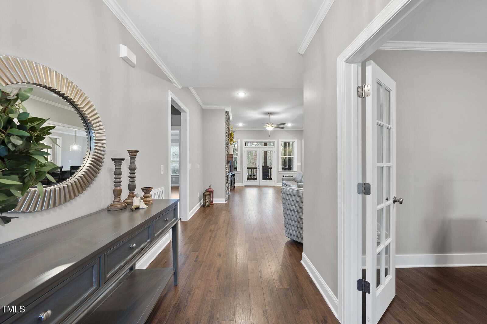 Hallway with hardwood floors, a console table with candles and a round mirror on the left, and a room with French doors on the right. The hallway leads to a living area with a ceiling fan and sofas.