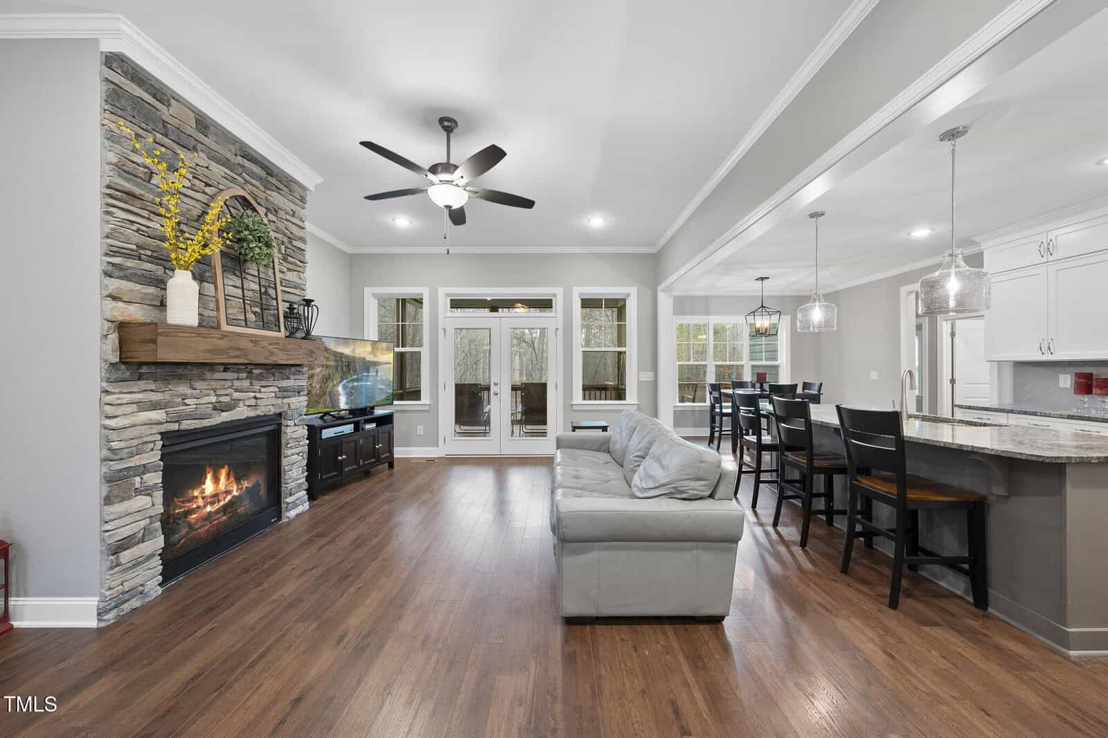 Open-concept living room and kitchen with wood floors, stone fireplace, gray sofa, ceiling fan, large windows, bar seating, and white cabinets. A dining table is visible near the windows; the fireplace has a wooden mantel.