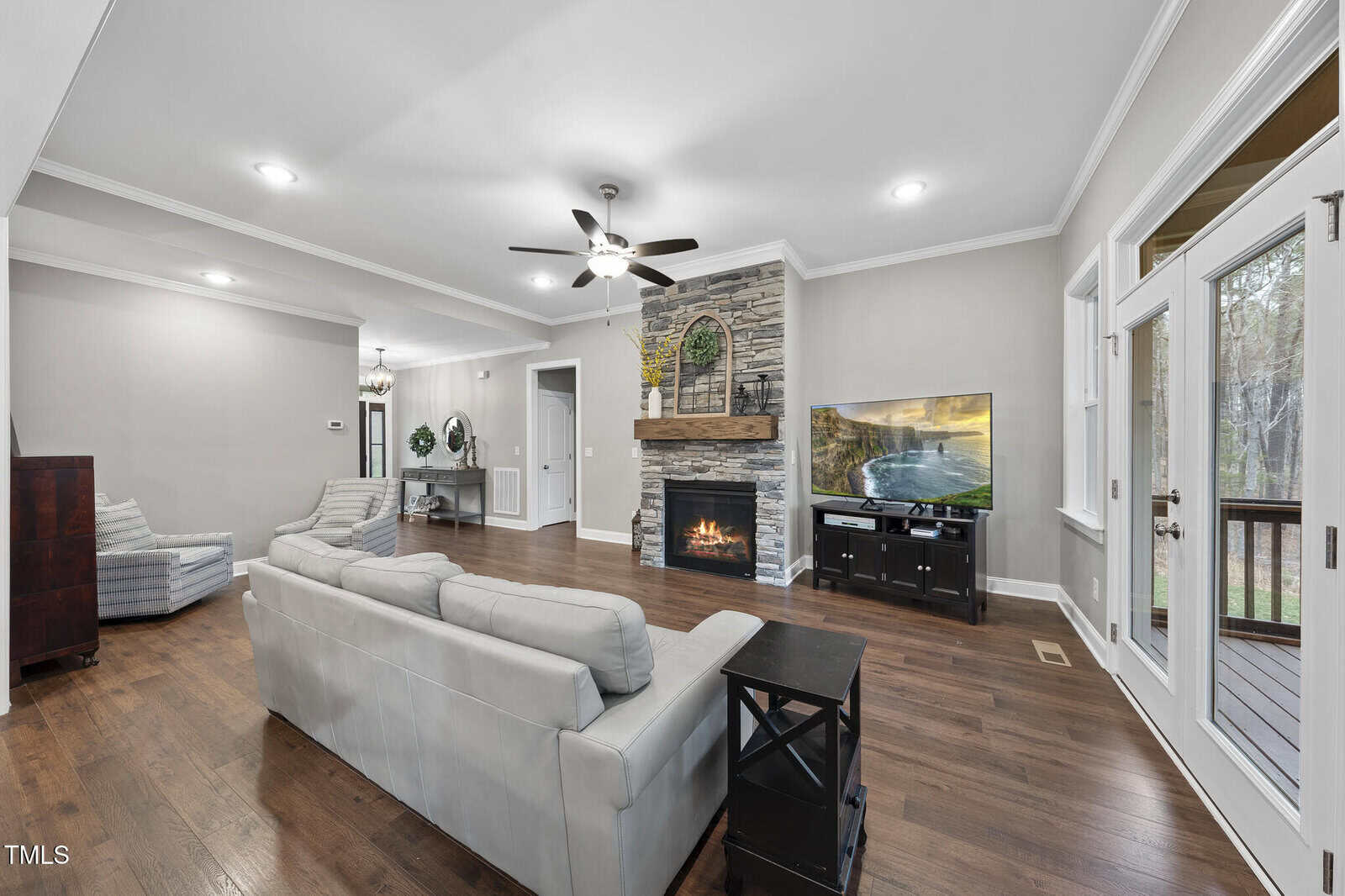 A living room with wood floors, light gray couches, a stone fireplace with a wooden mantel, a ceiling fan, a TV on a black stand, and glass doors leading outside. A hallway and armchair are visible in the background.
