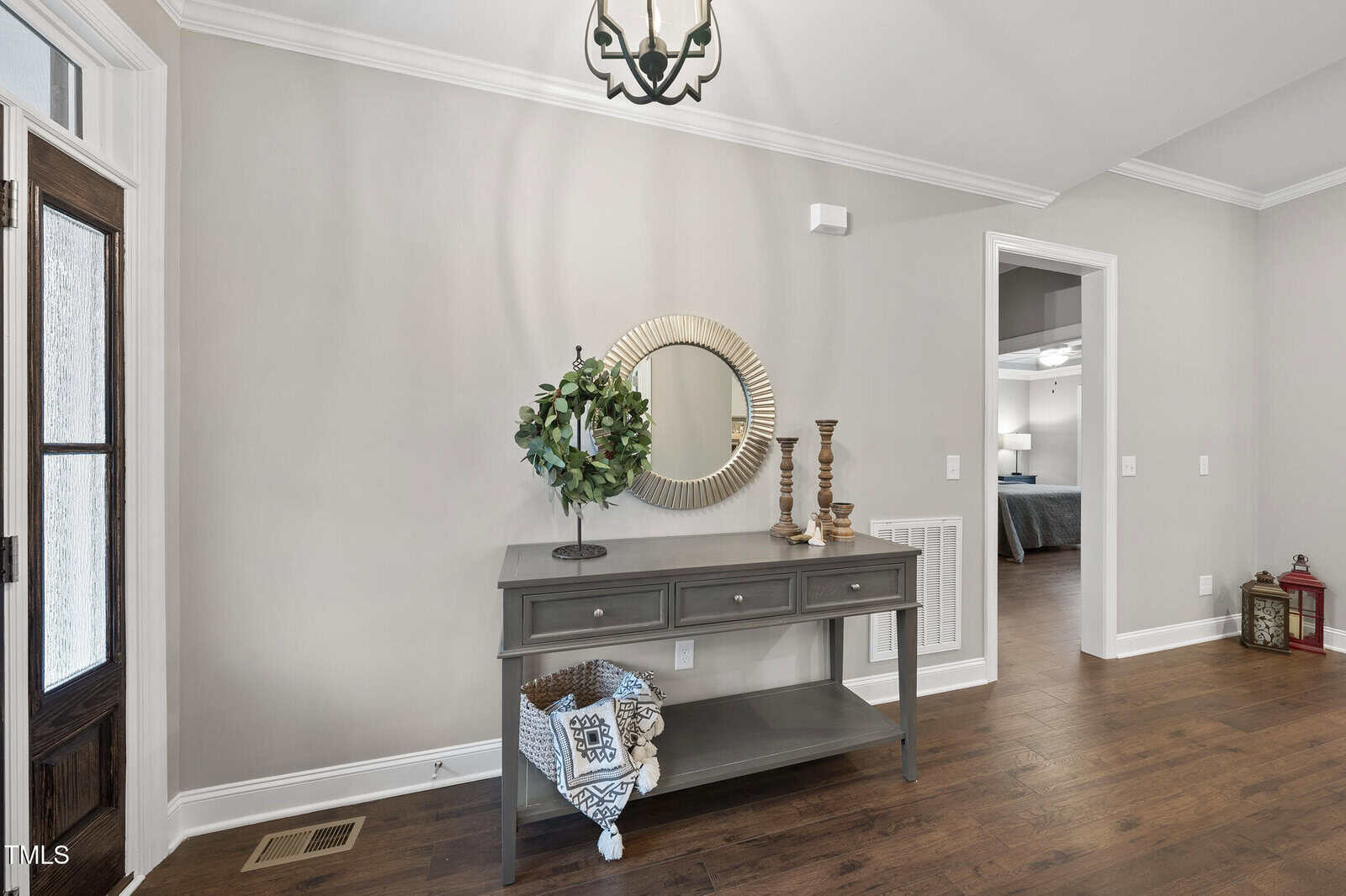 A hallway with wood flooring features a gray console table holding a round mirror, a potted plant, two candlesticks, and a decorative bowl. Decorative items and a folded blanket sit on the shelf below. An open doorway is visible.