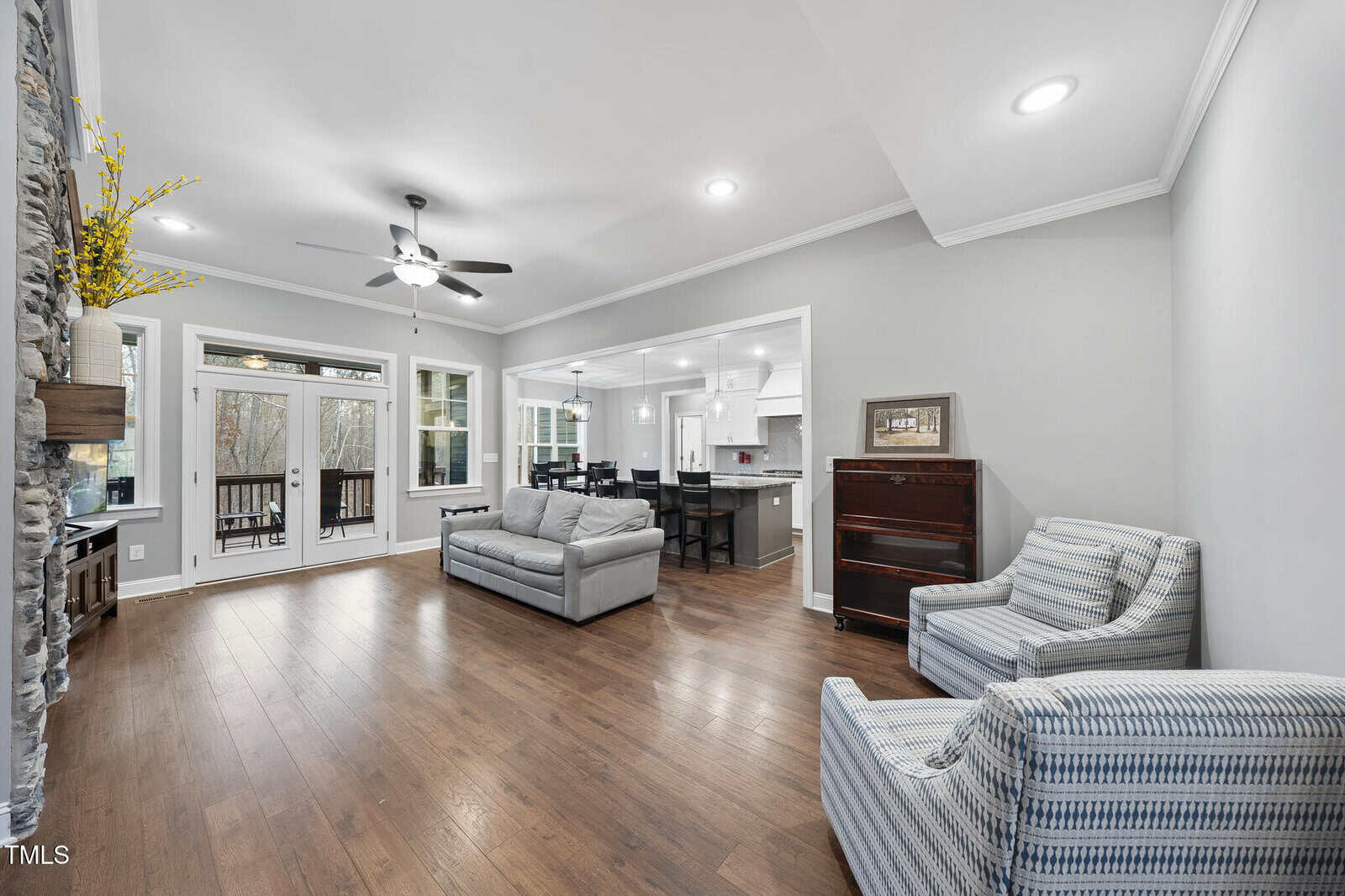 A spacious living room with wooden floors, gray walls, a sofa, two striped armchairs, a piano, and a stone fireplace. Large windows and glass doors lead to a covered porch, with an open view into the kitchen and dining area.