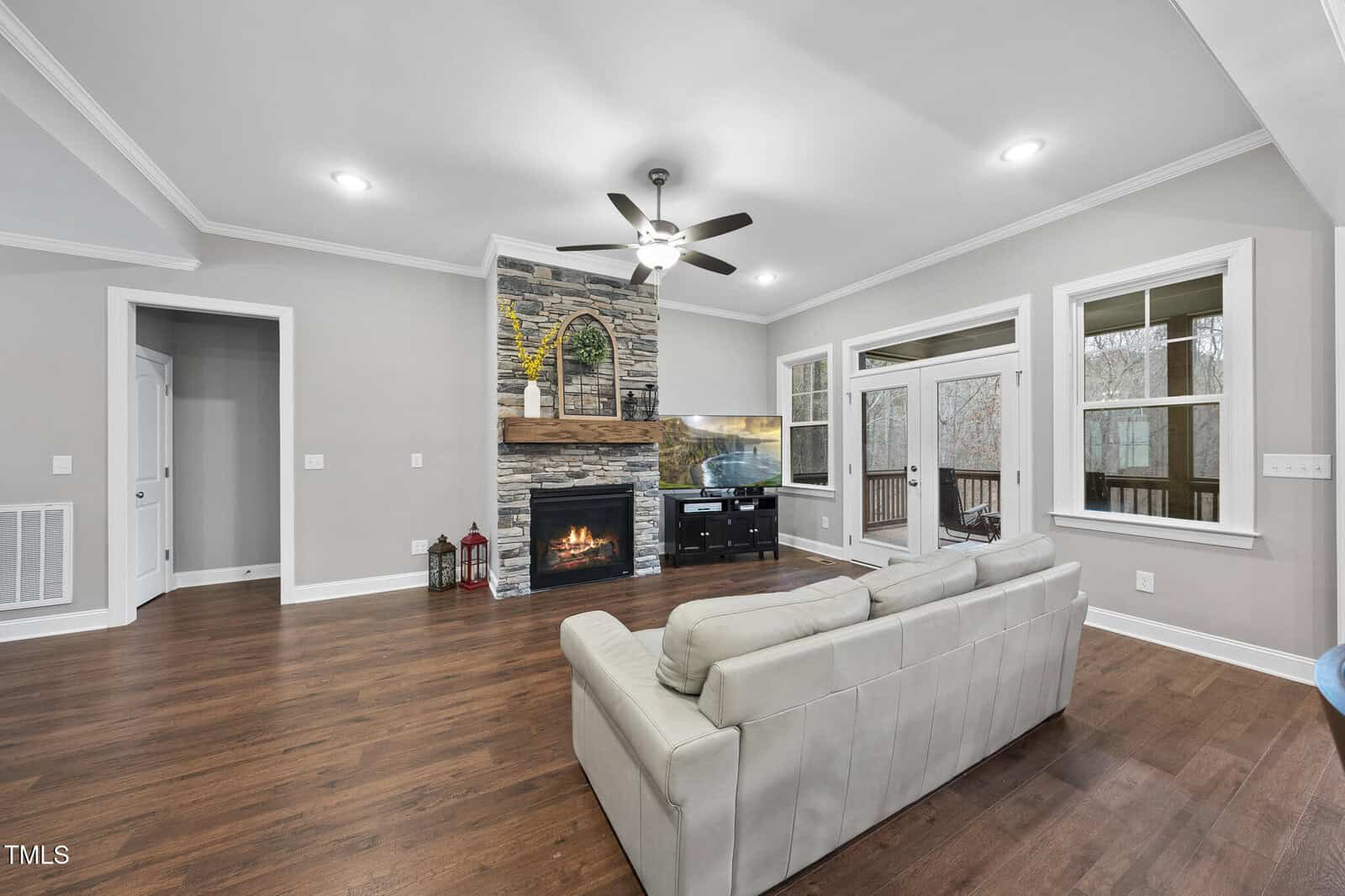 A living room with wood flooring, a white leather sofa, a stone fireplace with a fire burning, a TV on a stand, and large windows and doors leading to a deck. The walls are light gray with white trim.