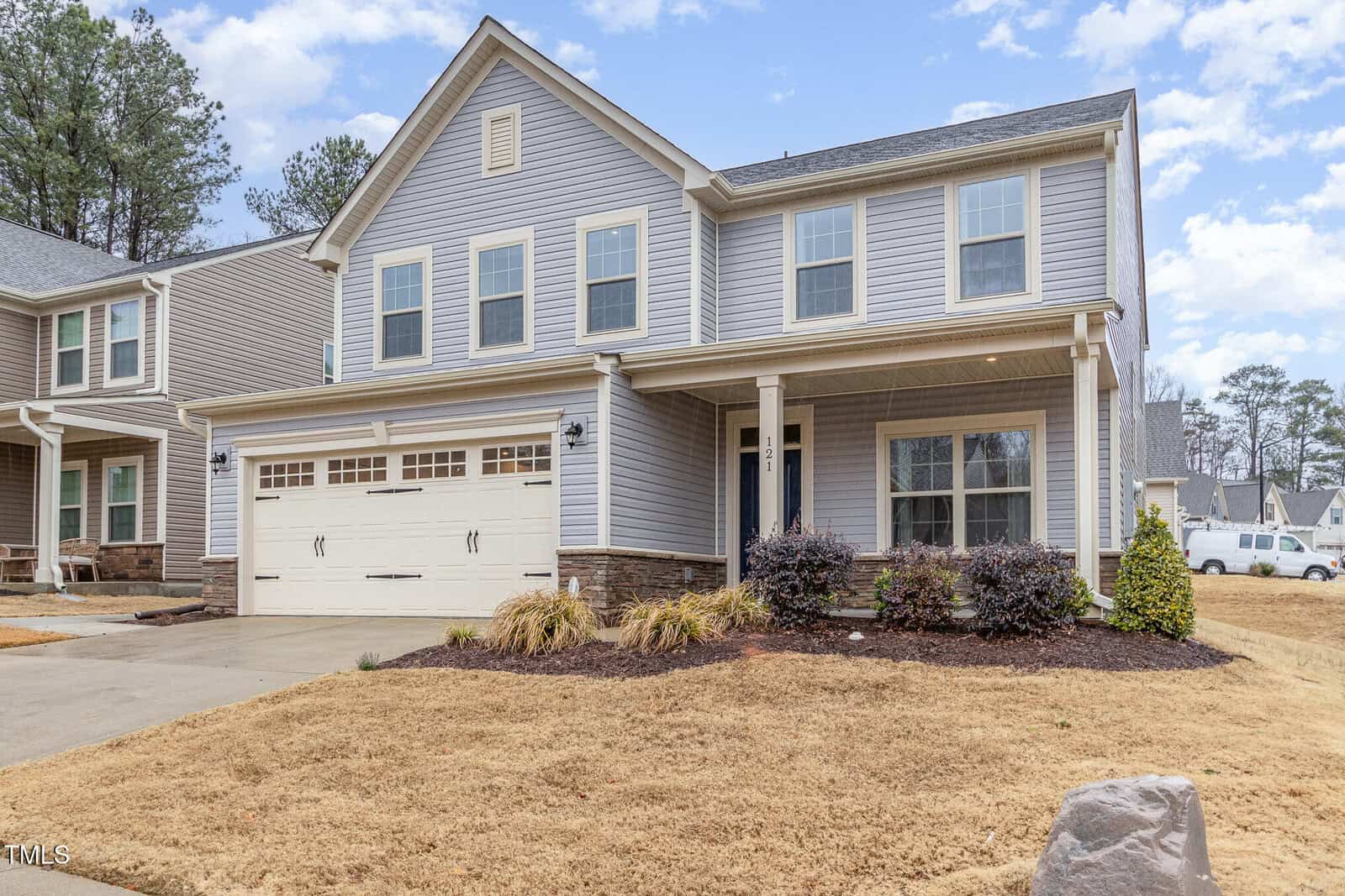 A two-story suburban house with gray siding, white trim, and a two-car garage. The front yard has dry grass, a small shrub garden, and a covered porch. Other houses and trees are visible in the background.