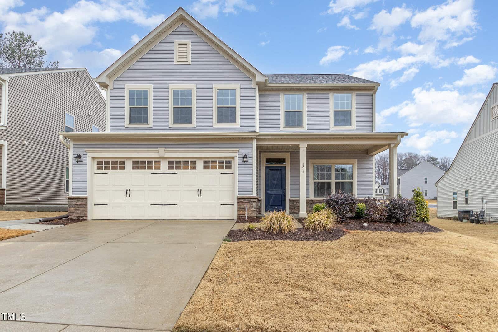 Two-story suburban house with grey siding, white trim, and a two-car garage. There is a covered front porch, driveway, and a lawn with some landscaping in front. The sky is partly cloudy.