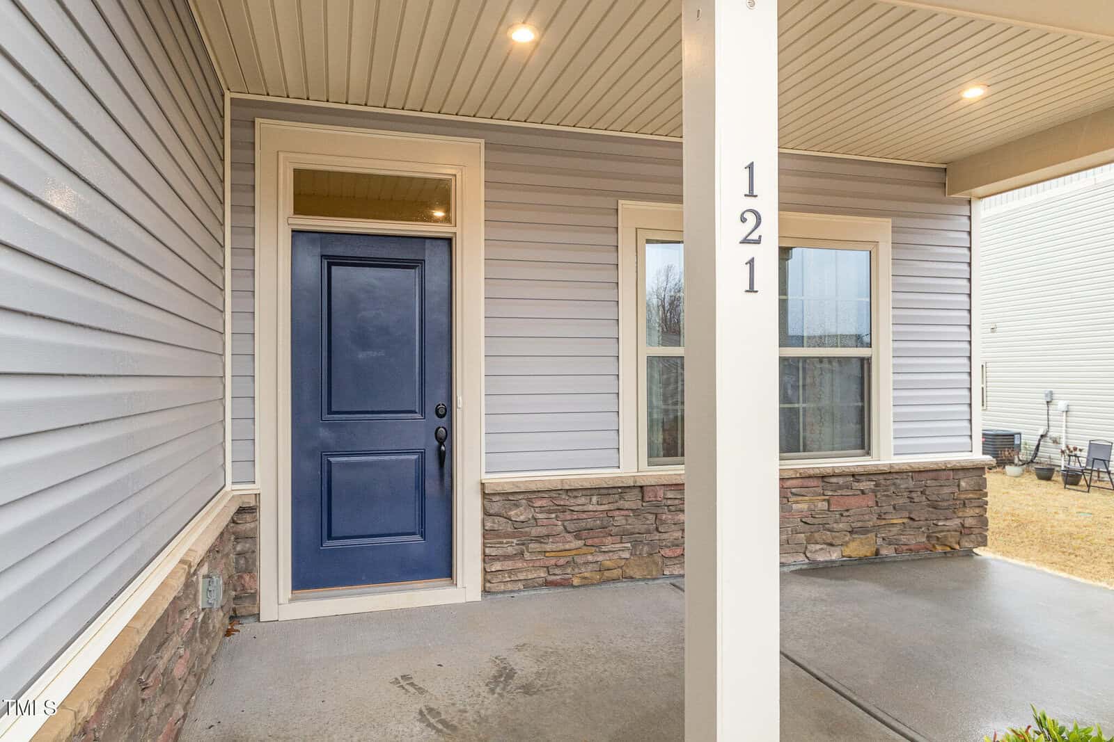 Front porch of a house with a blue front door, light gray siding, a partial stone facade, and the number 121 on a white post. Two windows are on the right side of the door. The concrete porch is partially wet.