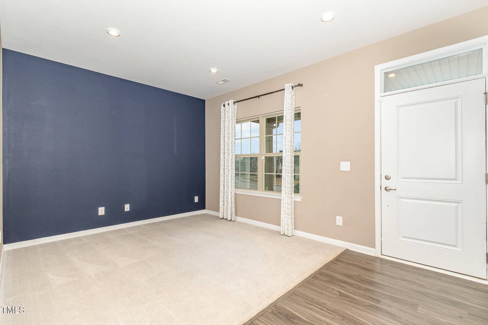 A clean, empty room with a beige carpet, wood flooring near the entrance, a navy blue accent wall, tan walls, a large window with curtains, and a white door with a window panel. Ceiling lights are recessed.
