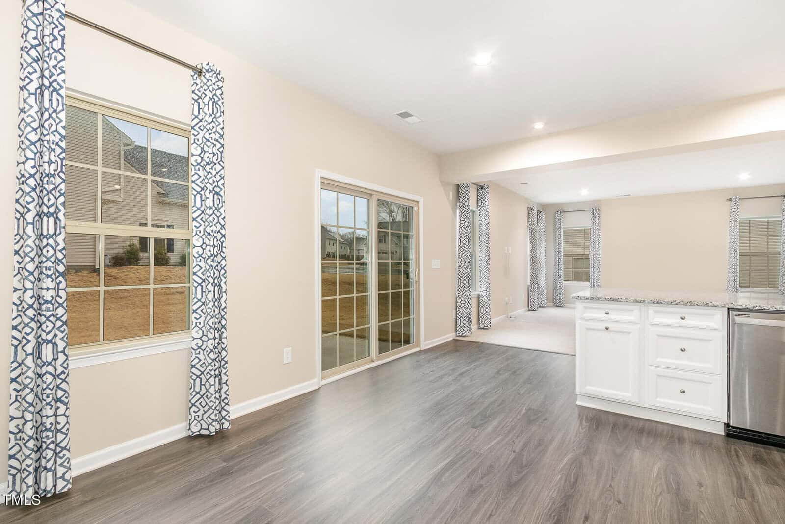An open-concept living space with light beige walls, wood-look flooring, white cabinetry, stainless steel appliances, large windows with patterned curtains, and sliding glass doors leading outside.