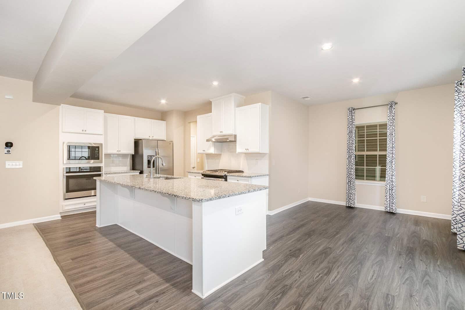 Modern kitchen with white cabinets, stainless steel appliances, a large island with granite countertops, and an open layout leading to a living area with wood-look flooring and large windows with patterned curtains.