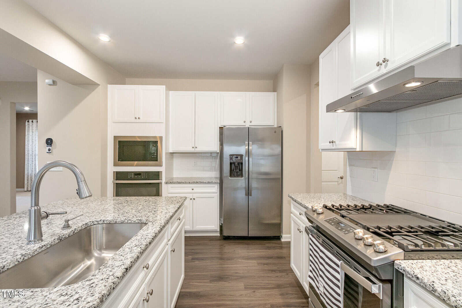 Modern kitchen with white cabinets, granite countertops, stainless steel refrigerator, built-in microwave and oven, gas stove with range hood, double sink, and wood flooring. Bright lighting and neutral wall color.