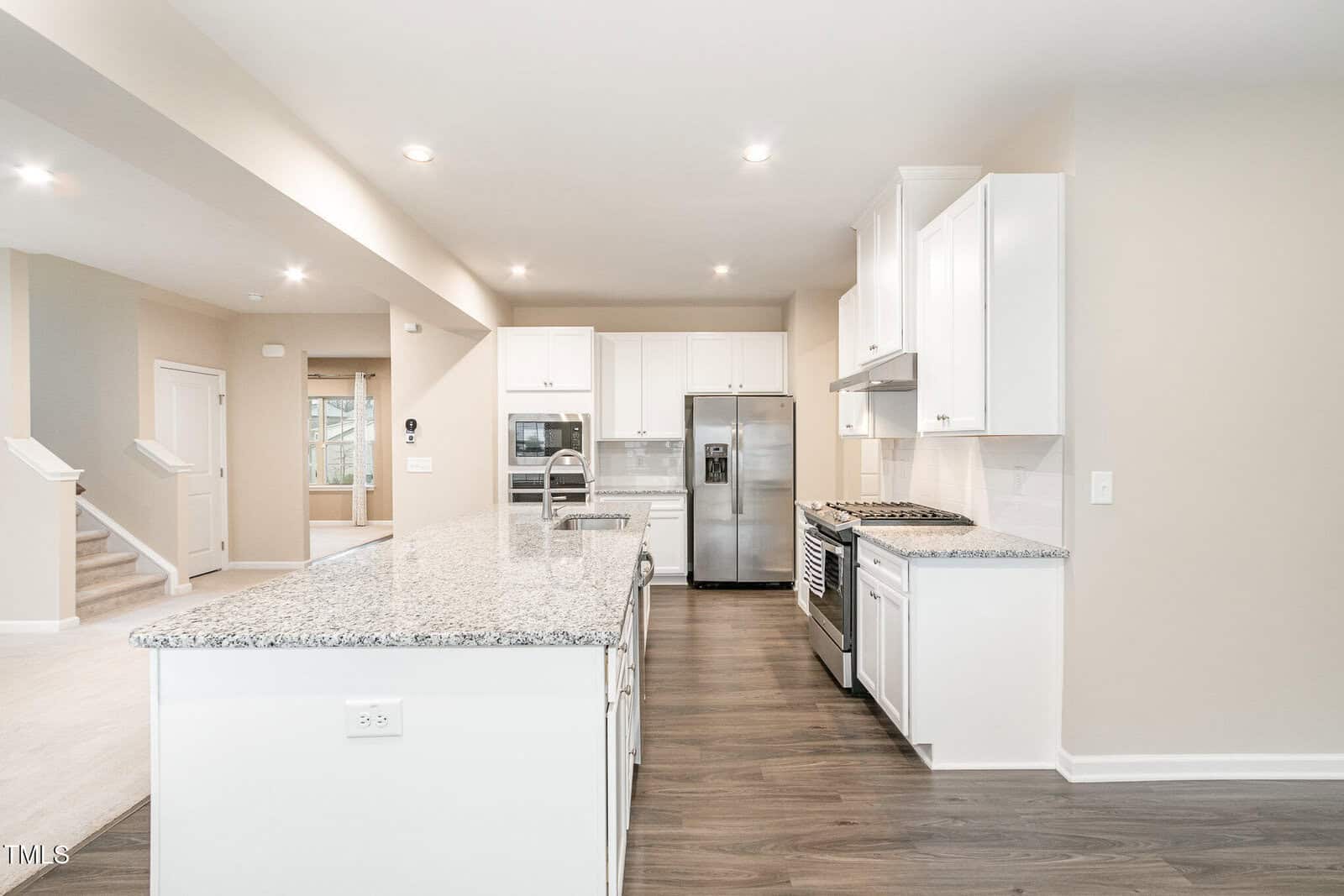 Modern kitchen with white cabinets, stainless steel appliances, granite countertops, and an island. Open layout leads to a living area with stairs visible on the left. Wood-style flooring and neutral walls throughout the space.