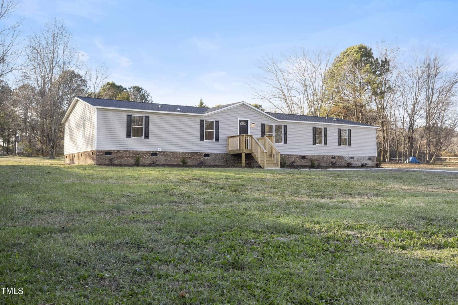 A single-story manufactured home with light-colored siding, black shutters, and a small wooden front porch, set on a brick foundation in a large grassy yard with trees in the background.