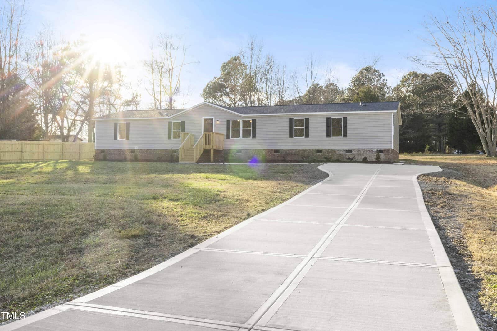 Single-story house with light-colored siding, brick foundation, and small front porch. The house is surrounded by grass and trees, with a long concrete driveway leading to the entrance. Sunlight is shining from the left.