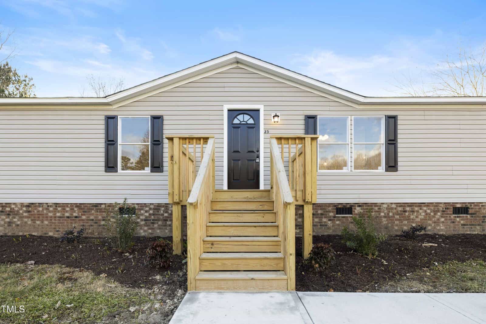 A single-story, beige manufactured home with black shutters, a black front door, wooden steps with railings, brick foundation, and minimal landscaping, set against a clear blue sky.