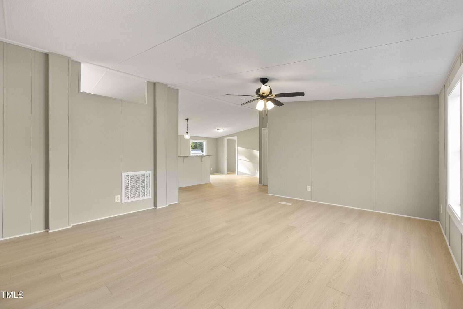 Spacious, empty living room with light wood flooring, beige walls, a ceiling fan with lights, and large windows letting in natural light. The room connects to an open dining area and kitchen.