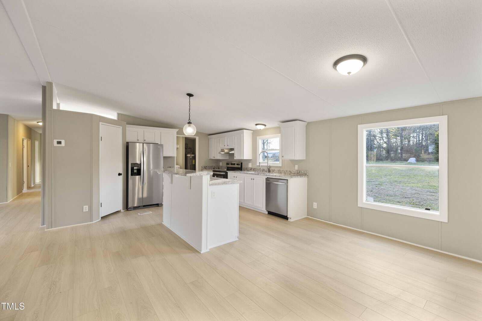Spacious kitchen and living area with light wood floors, white cabinets, stainless steel appliances, large window, and overhead lighting. Neutral walls and open layout with a hallway to the left.