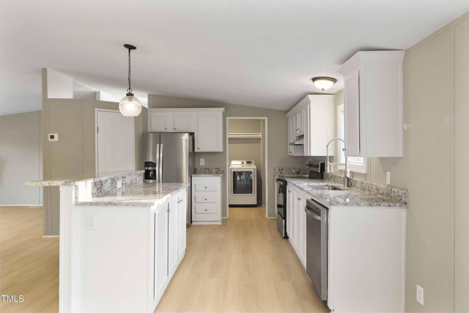 Modern kitchen with white cabinets, stainless steel appliances, marble countertops, light wood flooring, and a washer and dryer visible in a nook at the back. A pendant light hangs over the peninsula.