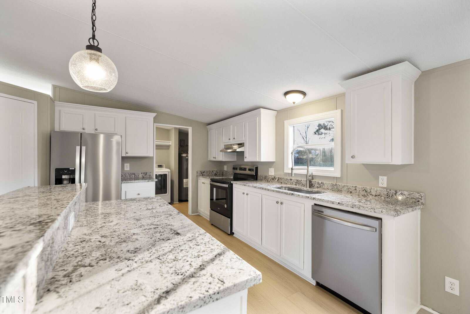Modern kitchen with white cabinets, granite countertops, stainless steel appliances, a window over the sink, a hanging light fixture, and an open layout with light wood flooring.