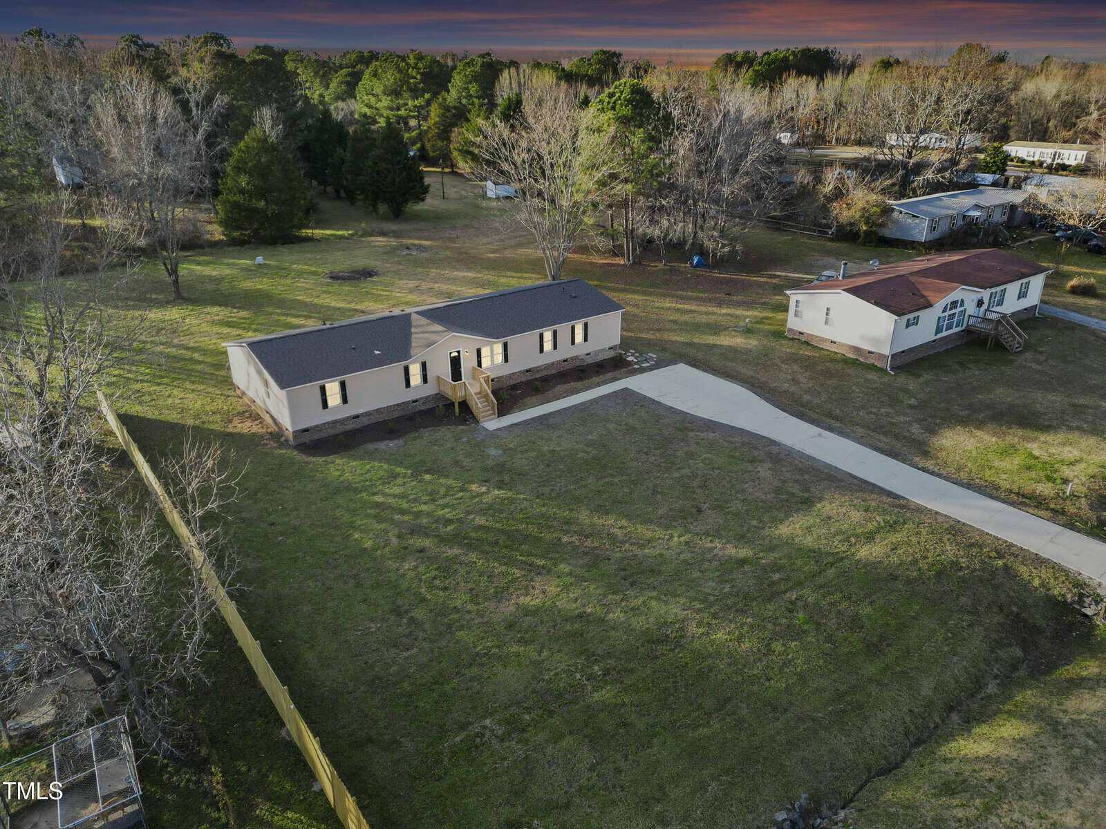 Aerial view of a beige single-story manufactured home with a front porch, large yard, long driveway, and a neighboring similar house, surrounded by trees and grass under a dramatic evening sky.