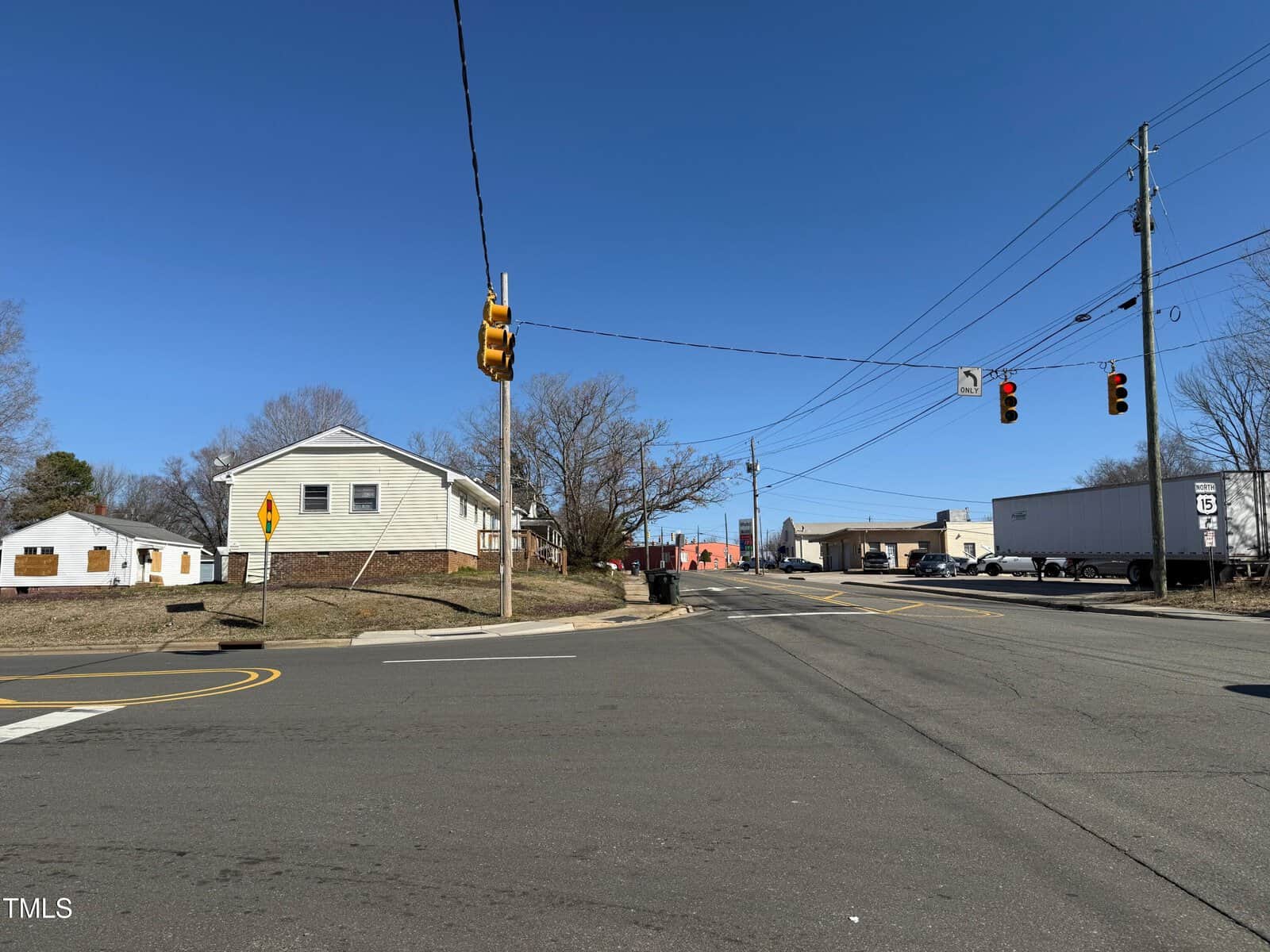 An intersection with traffic lights on a clear day. A two-story house is on the left, and an industrial building with a truck is on the right. Power lines and a road sign are visible. The sky is clear and blue.