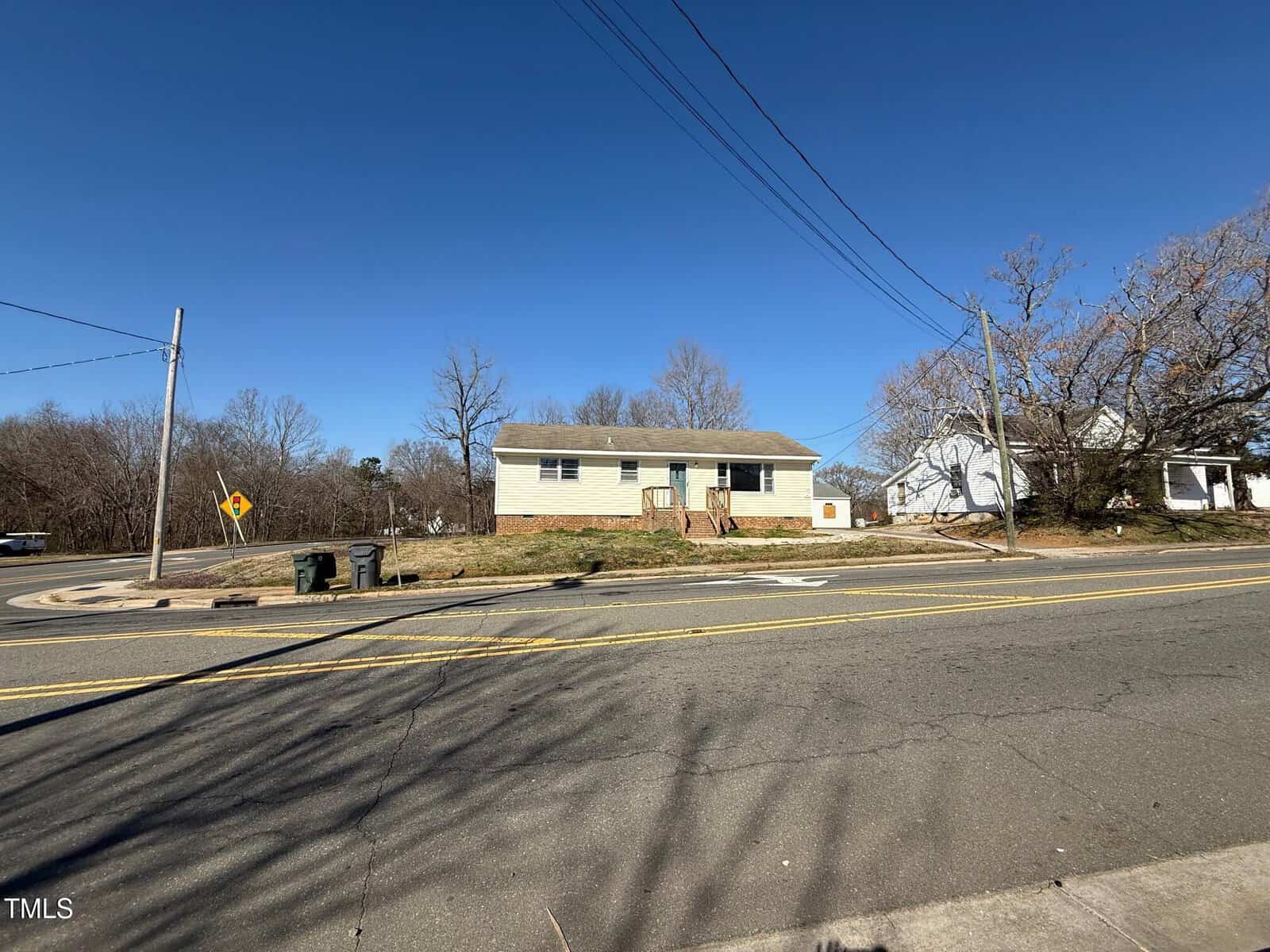 A single-story beige house with a gray roof is situated on a corner lot. The house is bordered by a road with double yellow lines. Leafless trees surround the property, and utility lines cross the clear blue sky.