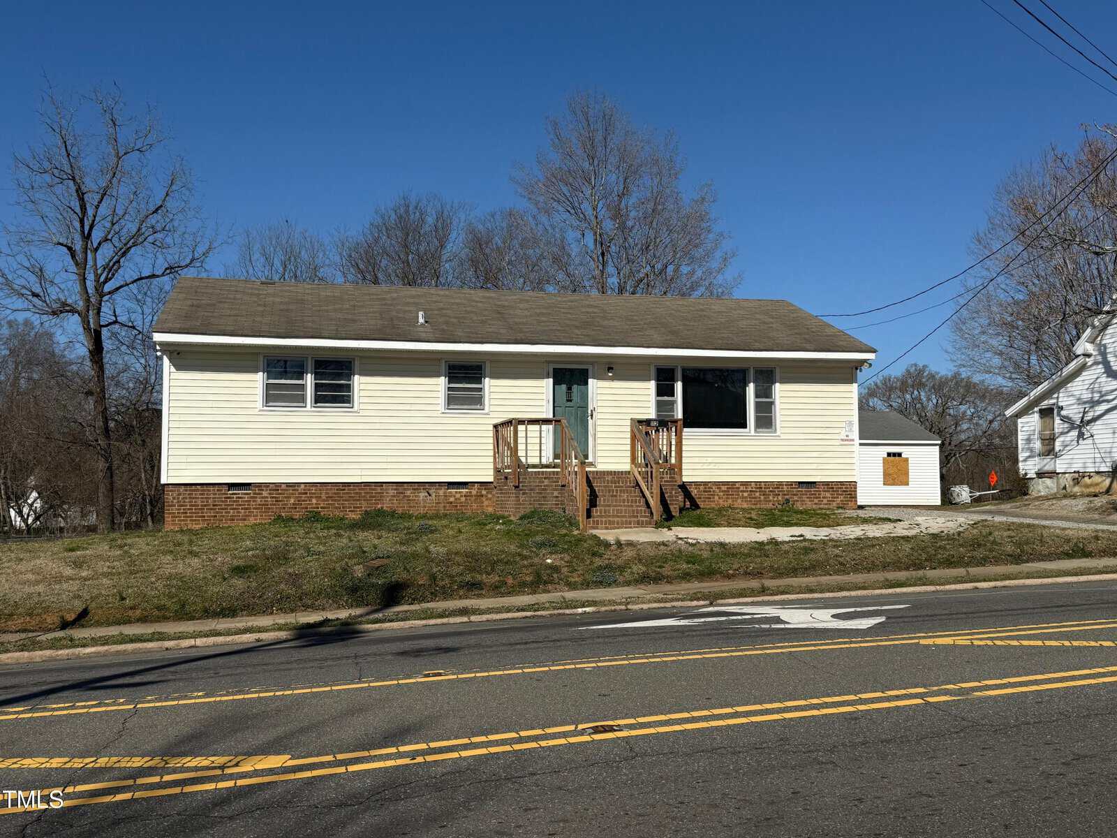 A single-story, beige house with a gray roof and white trim sits behind a two-lane road. The house has a front porch with stairs and railing. Sparse grass and leafless trees are visible in the yard and surroundings under a clear blue sky.
