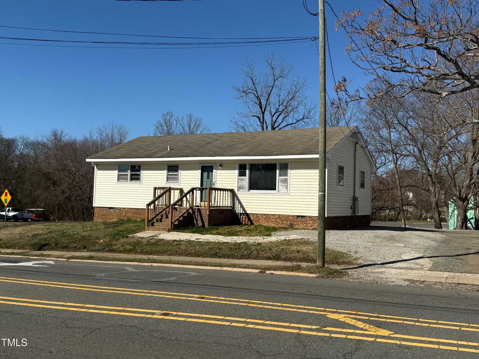 A single-story house with a light-colored exterior and an asphalt shingle roof is situated near a road with double yellow lines. The house features a small front porch with wooden steps and a railing. Leafless trees are visible in the background.