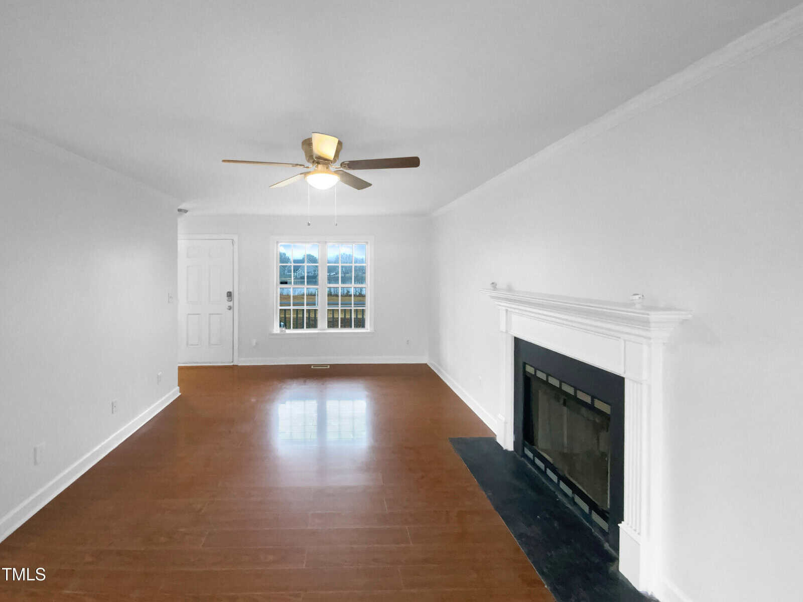 A bright living room with white walls, wood flooring, a ceiling fan with light, a white fireplace with black trim, and a window beside a white front door. The room is empty and well-lit by natural light.