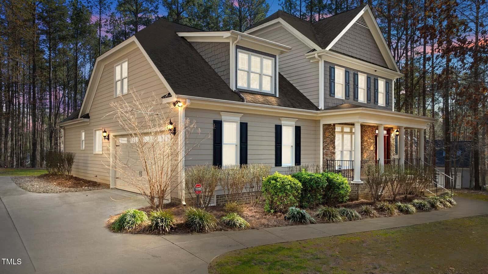 A two-story house at 1196 Smith Creek Way, with beige siding, white trim, and dark shutters is surrounded by landscaped bushes and a lawn. Located in Wake Forest, it boasts a gabled roof and a porch. The backdrop features trees against a sky transitioning from blue to pink.