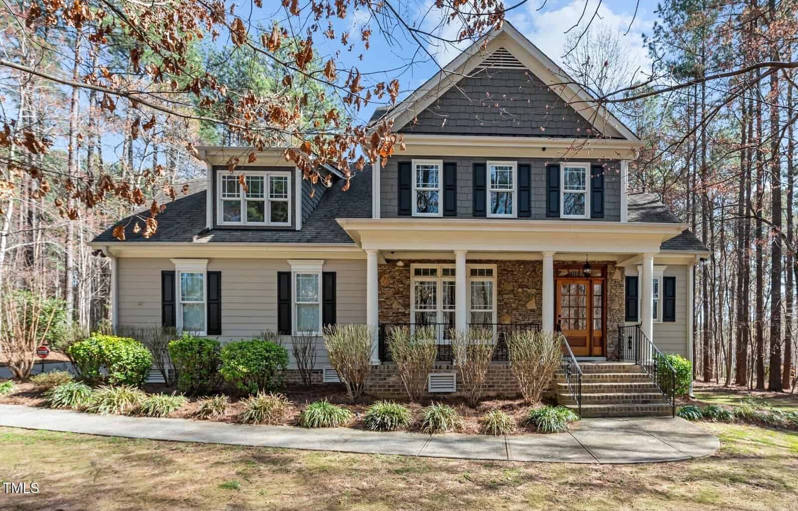 Two-story house with gray siding, white trim, and dark shutters. It features a covered front porch with white columns and steps leading to a wooden door. The yard has shrubs along the walkway and tall trees in the background.