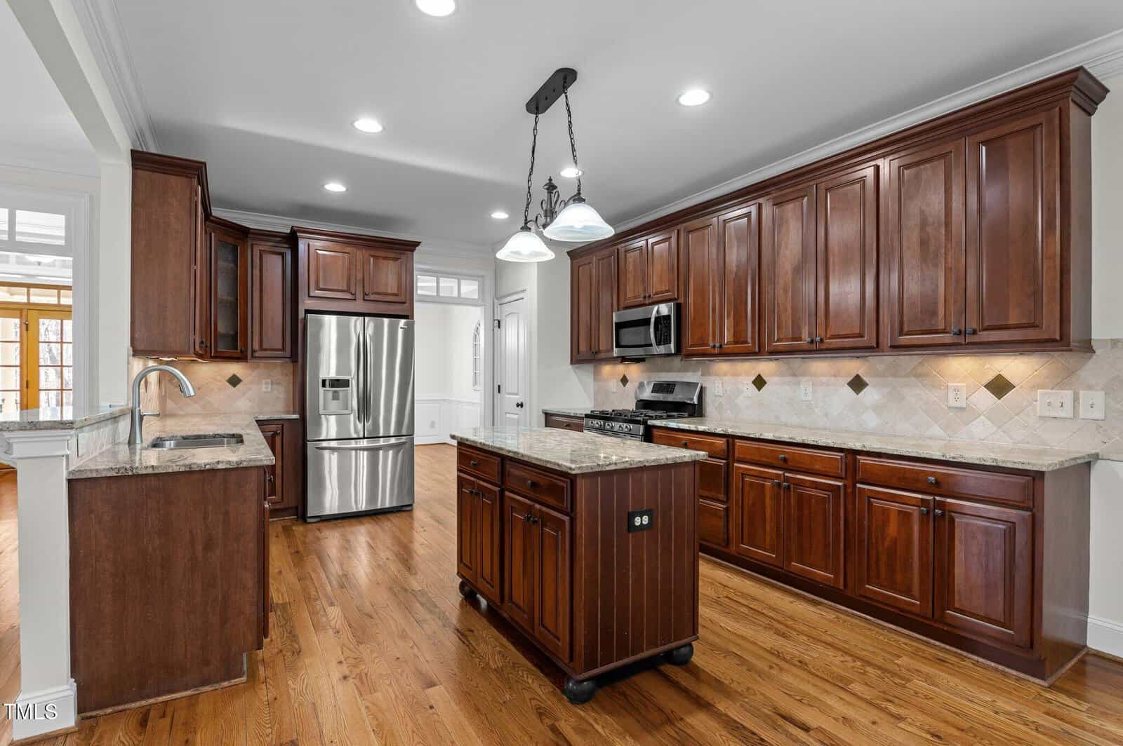 A spacious kitchen with wooden cabinets, a stainless steel refrigerator, oven, and microwave. An island with a sink stands in the center. The flooring is wood, and the ceiling features recessed and pendant lighting. Walls have light, neutral tiles.