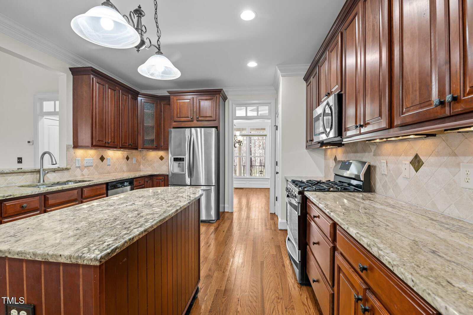 A modern kitchen with dark wood cabinets and stainless steel appliances. There is a central island with a granite countertop, a gas stove, and a large refrigerator. The backsplash features beige tiles, and recessed lighting is present.