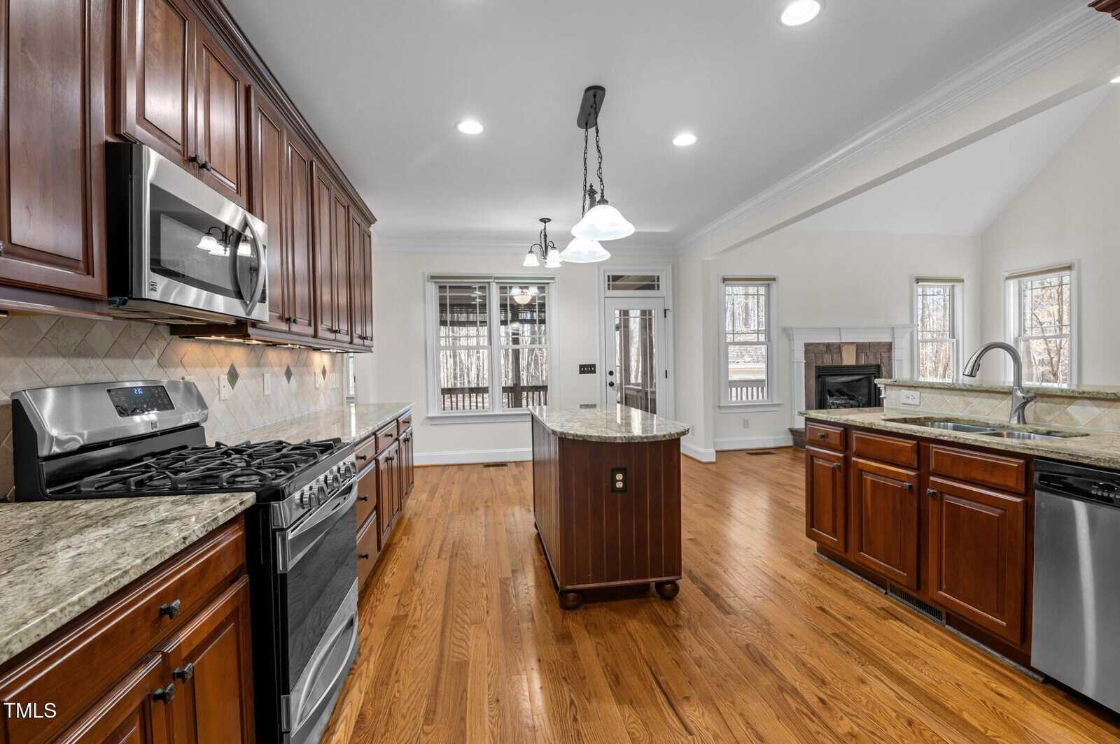 A spacious kitchen with wooden cabinets and a marble island. It features stainless steel appliances including a stove, microwave, and dishwasher. The room has hardwood floors, recessed lighting, and large windows providing ample natural light.
