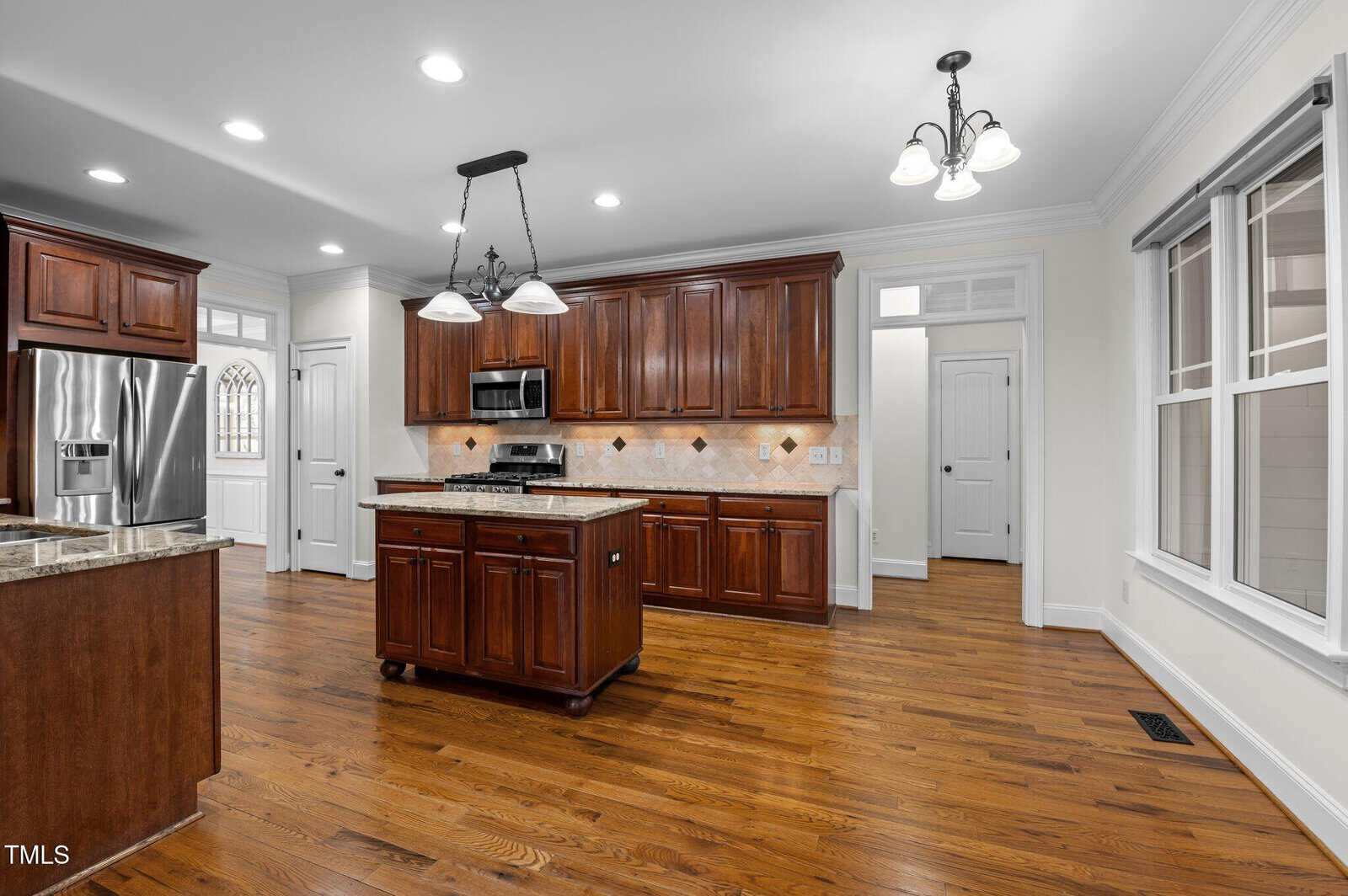 A modern kitchen with wooden cabinetry, stainless steel refrigerator, oven, and microwave. A central island features a granite countertop. The room has wooden flooring, recessed lighting, and a window. A door leads to a separate area.