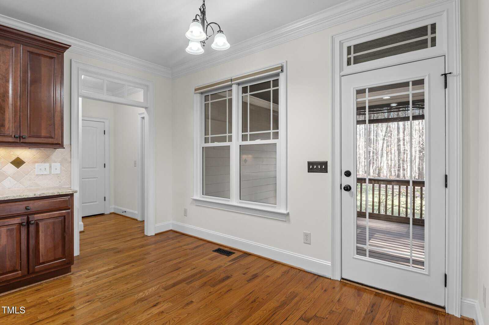 Kitchen area with hardwood floors, white walls, and a ceiling light fixture. A wooden cabinet is partially visible on the left. There is a window and glass door leading to a deck outside, with trees in the background.